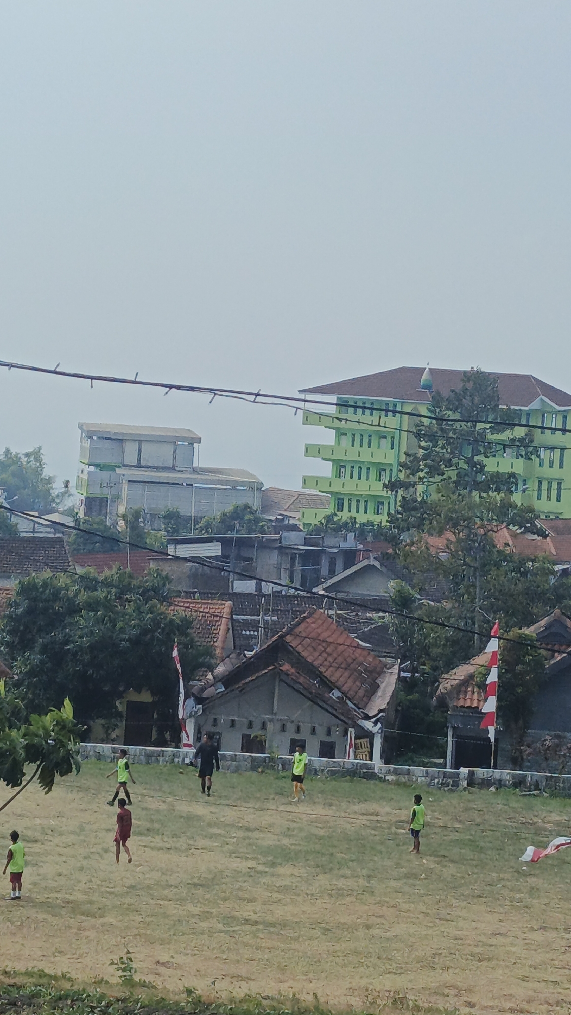 A group of boys are playing soccer on a dry, grassy field. The field is surrounded by a low stone wall. In the background, there are houses with red tile roofs and a large, multi-story green building with a dome. There are trees and electrical wires in the foreground and background. A red and white flag is visible on a pole. The sky is overcast. The scene appears to be in a village in Sajen, Indonesia.