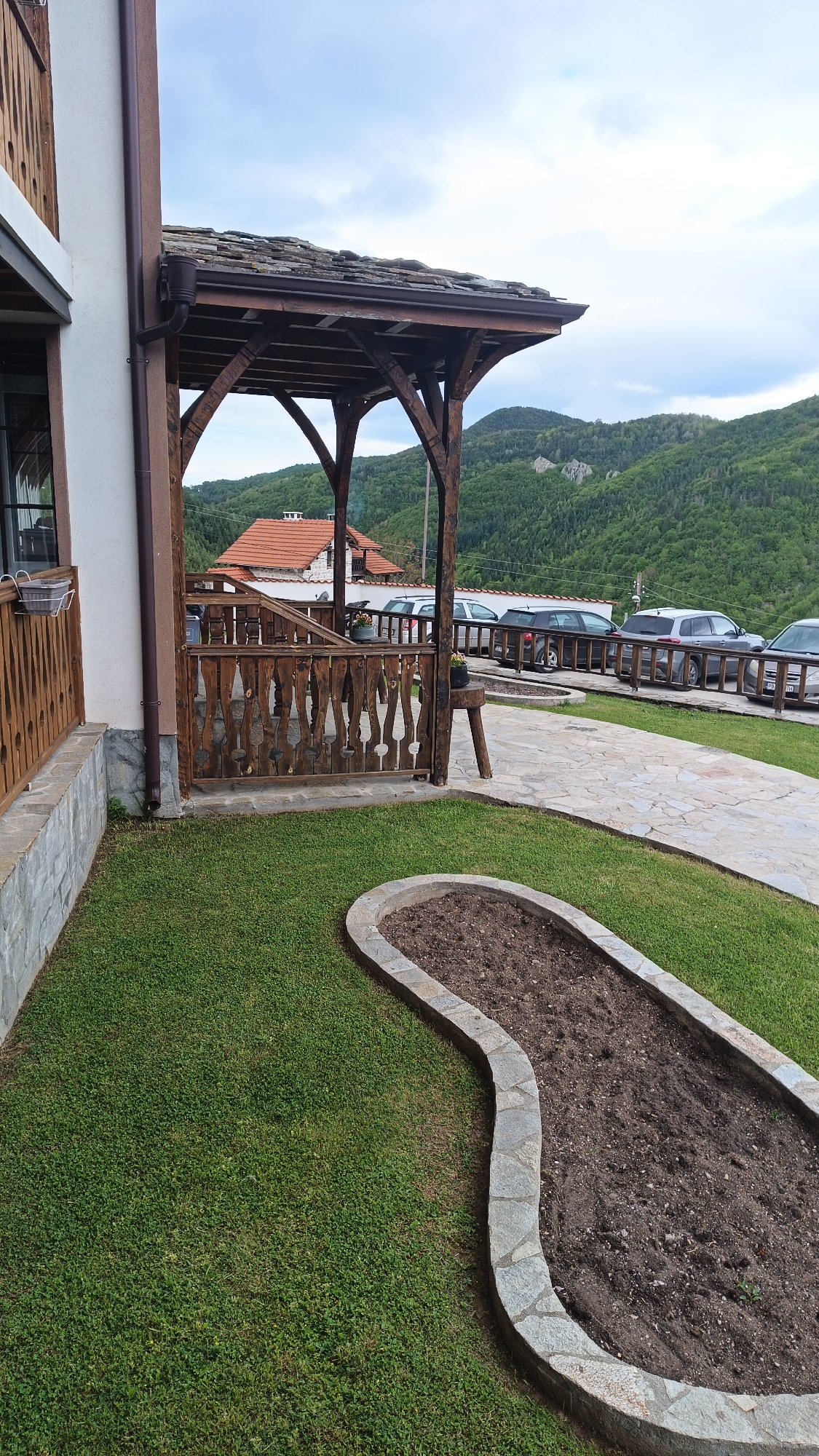 The image shows a landscaped area near a building, possibly a residence, in Vrata, Bulgaria. In the foreground is a curved garden bed filled with dark soil and bordered with gray stone. Beyond the bed is a green lawn. To the left, the corner of a building is visible with a wooden porch and stone foundation. The porch structure has wooden railings and posts, topped with a tiled roof.
In the background, across a stone-paved path, several parked cars are lined up alongside a wooden fence. Beyond the cars and fence are rolling hills covered with dense green vegetation, suggesting a mountainous or forested region. The sky is overcast with hints of blue.
The scene appears serene, presenting a well-maintained property in a rural setting. There are no visible people or interactions.