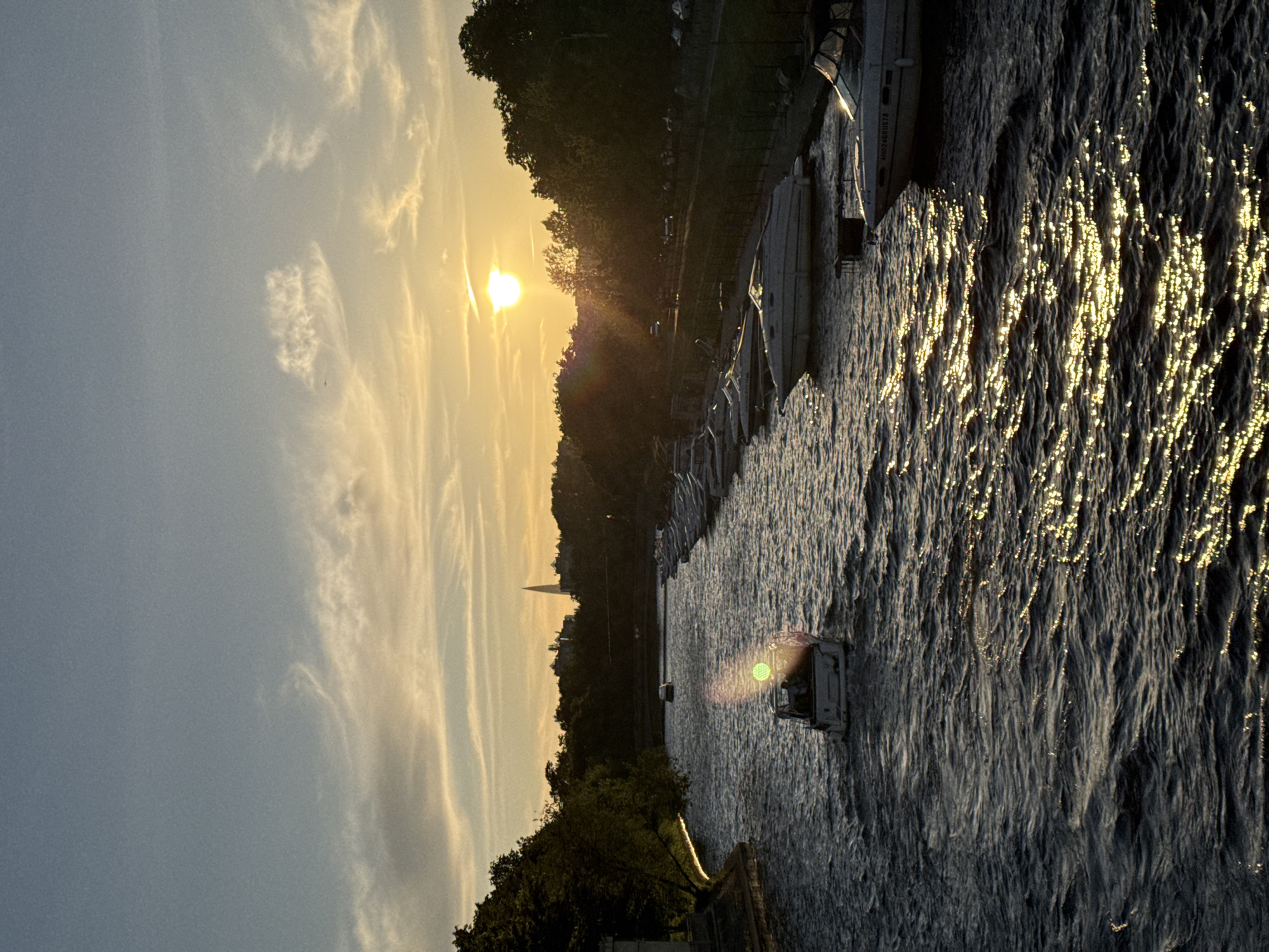The image depicts a sunset over the Malaya Neva River in Saint Petersburg, Russia. The sun is low on the horizon, casting a warm golden light across the water and the sky. Several boats are moored along the right bank of the river, and another boat is visible in the middle of the river, moving away from the viewer. The left bank is lined with trees, and in the background, several buildings are visible, including a tall, spire-like structure. The water is choppy, with sunlight reflecting off the ripples. The overall atmosphere is serene and picturesque.