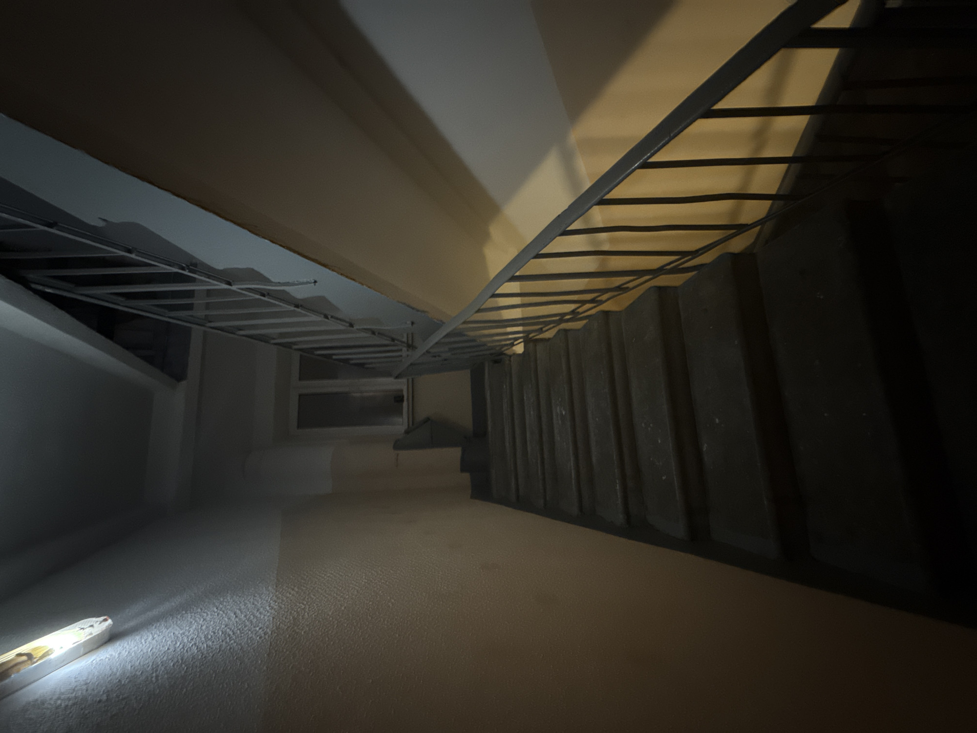 The image depicts a dimly lit stairwell, likely indoors, with metal railings on both sides. The lighting source, possibly a flashlight or work light, is positioned at the bottom left corner, casting a strong beam of light across the floor. The stairwell's construction includes what appears to be concrete or painted risers and treads, with a textured surface on the floor.
Given the prompt's reference to Mytishchi, Russia, it's plausible that this stairwell is located within a residential building, commercial structure, or industrial facility in that city. However, without additional context or visual cues, it is impossible to definitively pinpoint the specific location within Mytishchi. No people are visible in the image. The subjects present are the stairs and railings. The actions are unknown as the scene is static. There are no explicit interactions. The environment is dark and seemingly indoors.