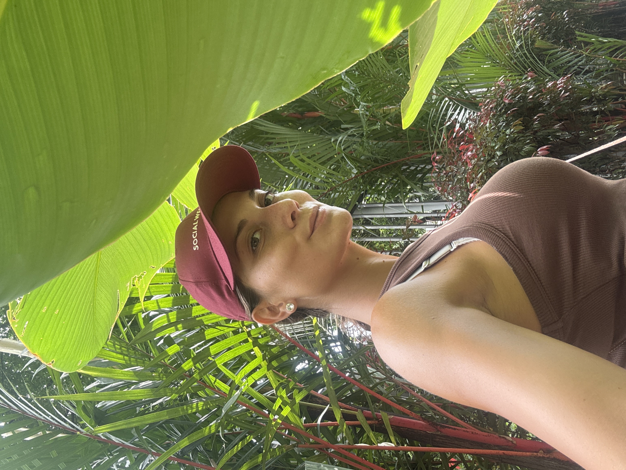 A woman wearing a brown tank top and a burgundy baseball cap is standing in a tropical garden. The cap has the word "SOCIAL" written on it. She is looking upwards towards the large green leaves above her, seemingly shading her. The background is filled with various lush green plants and some red-stemmed flora. The location appears to be a garden within Kuala Lumpur, Malaysia, given the tropical vegetation. The angle of the photo suggests it was taken from a lower perspective.
