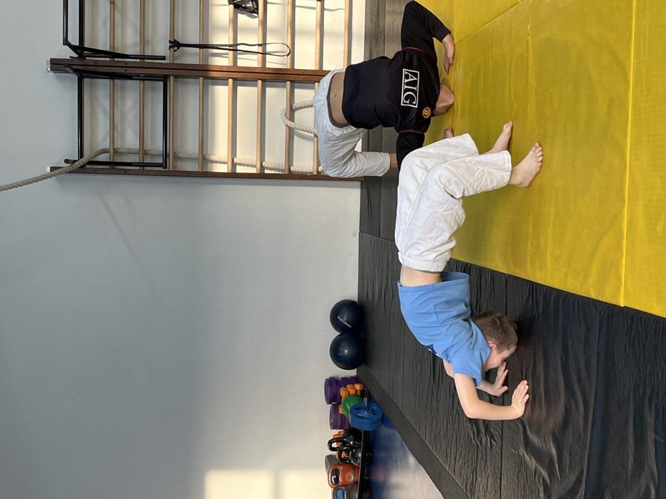 Two young males are depicted indoors within a gym or training facility in Krasnoyarsk, Russia. Both individuals are performing a 'bridge' or 'wheel' flexibility pose on contrasting athletic mats. The male on the left, wearing a light blue t-shirt and white athletic pants, is positioned on a dark grey/black mat with his hands and the crown of his head contacting the surface, body arched upward. The male on the right, wearing a black long-sleeved athletic shirt bearing an 'AIG' logo and white athletic pants, is similarly inverted on a bright yellow mat, hands and head on the surface, supporting his arched body. Both are barefoot. The background on the left reveals a white wall adjacent to assorted gym equipment, specifically black and grey kettlebells, stacked purple, blue, and orange weight plates, and two dark blue medicine balls. To the right, a wooden wall ladder (Swedish ladder) is visible, from which a thick white rope hangs vertically. A rectangular shadow on the upper left wall indicates natural light entering the space. The scene is brightly illuminated.