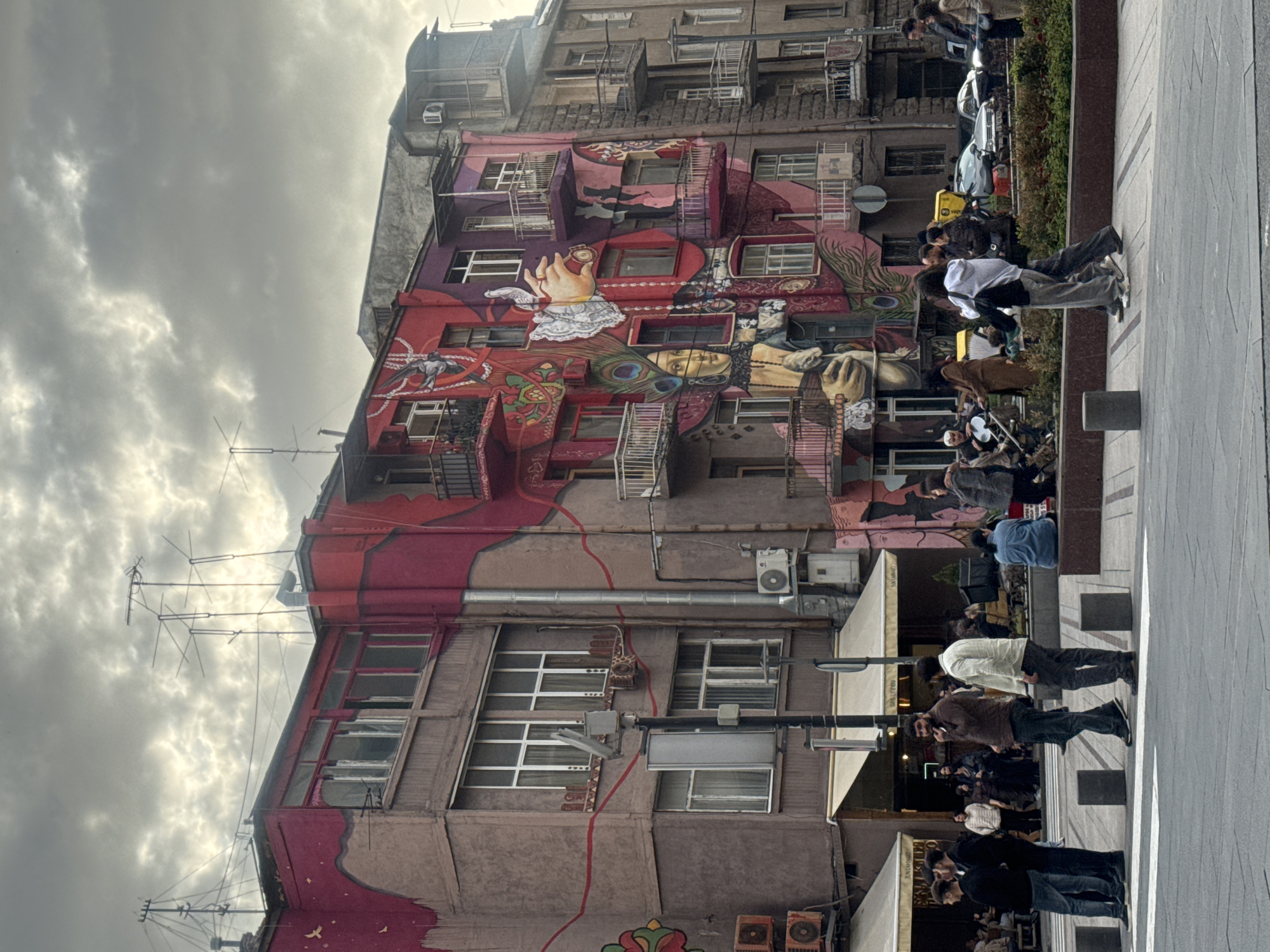 The image shows a multi-story building in Yerevan, Armenia, distinguished by its colorful mural that spans several floors. The mural features artistic representations of faces, hands, and decorative elements such as peacock feathers. The building itself is of aged construction with visible architectural details like balconies, windows of varying sizes, air conditioning units, and external conduits. There are television antennas atop the building.
The environment includes a paved pedestrian area adjacent to the building. Pedestrians are seen on the sidewalk, some appearing to be tourists or locals observing the mural. A series of awnings suggest the presence of businesses or restaurants lining the street. The sky is overcast with heavy clouds.