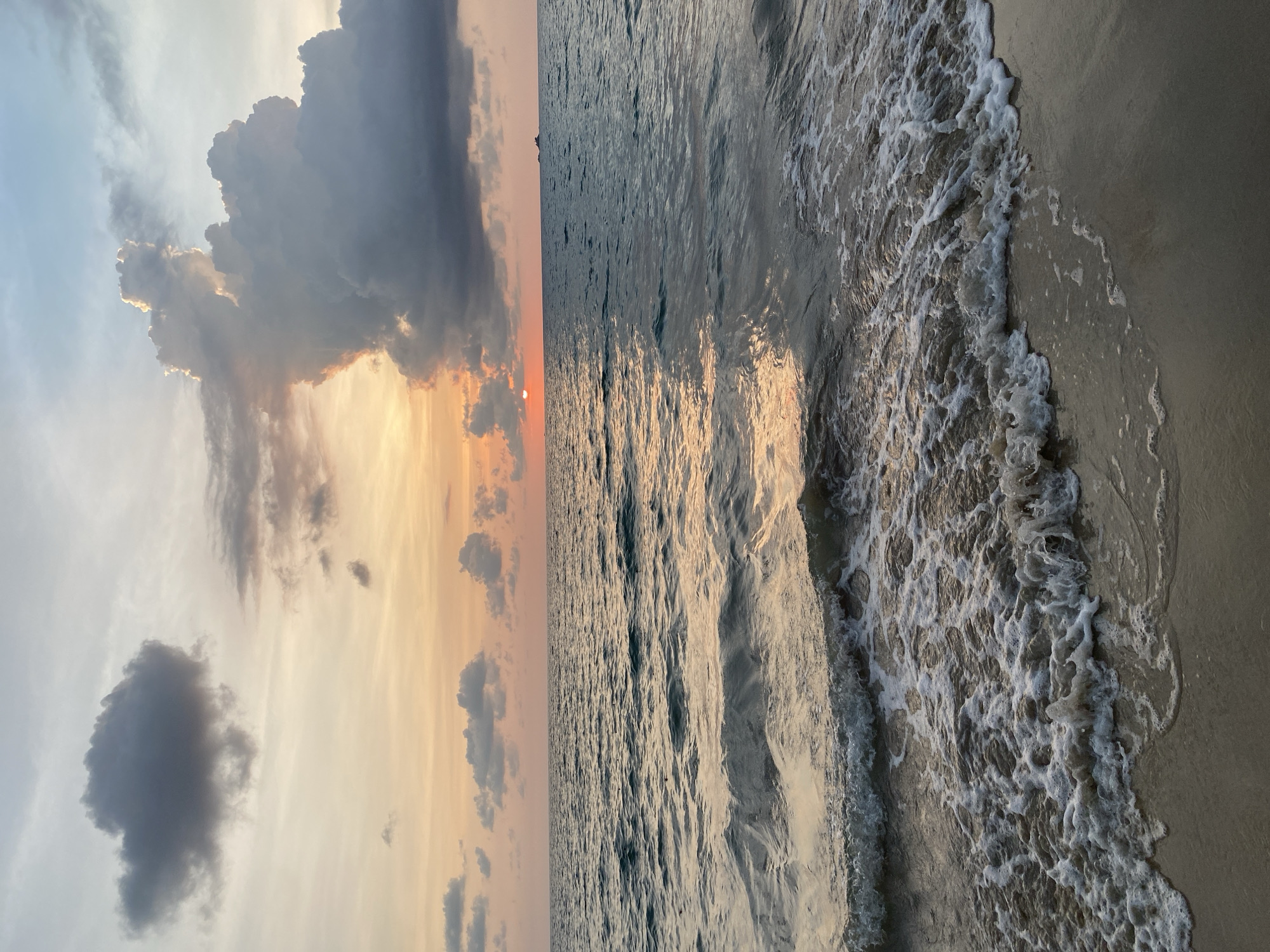 A scenic coastal landscape is depicted at either sunrise or sunset, with the horizon line dividing the sky and the sea. The sun, a small orange orb, is partially visible and obscured by large, dark, and irregularly shaped clouds near the horizon, casting warm hues of orange, pink, and yellow across the lower sky. Above, the sky transitions to lighter blues and grays, with scattered, smaller clouds.
The ocean occupies the central part of the frame, showing gentle waves with visible ripples reflecting the warm light from the sun. The water closest to the shore forms breaking waves, characterized by white, frothy foam that contrasts with the darker water. These foamy waves gently lap onto the sandy beach, which occupies the foreground of the image. The sand appears wet and smooth where the waves recede. The overall atmosphere is peaceful and serene, showcasing natural beauty. There is no visible text in the image. The scene is consistent with a tropical beach setting, such as Choeng Thale, Thailand.