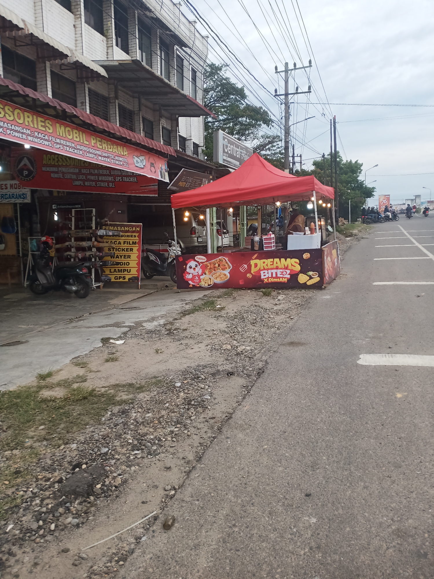 In Lhokseumawe, Indonesia, a street scene unfolds with a car accessory shop on the left, its red awning displaying "AKSESORIES MOBIL PERDANA" and listing services like window film, audio, and GPS trackers. Adjacent to it, a food stall with a red canopy operates, displaying a banner for "DREAMS BITE x DimsAM" featuring images of fried chicken and dim sum. Several motorcycles are parked near the shop and food stall. The street itself is asphalt, with a gravel and dirt shoulder. In the background, utility poles and wires crisscross the overcast sky, and several people on motorcycles are visible further down the road.
