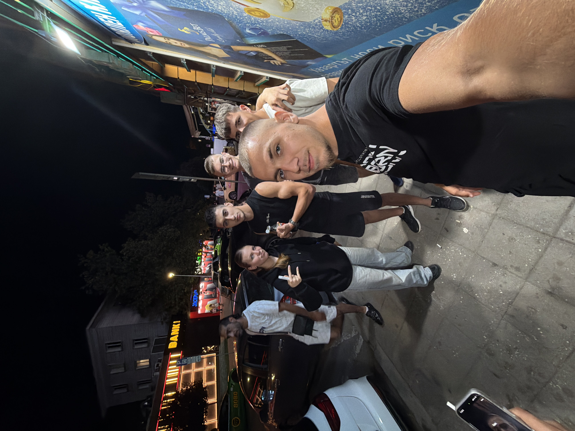 In Ruse, Bulgaria, at night, a group of five young adults pose for a selfie on a sidewalk. The group consists of three males and two females. The building behind them features neon signage for "BANK!" and "JACKPOTS," indicating a commercial area. Several cars are parked on the street. A large blue advertisement poster is visible on the right side of the image. The ground is paved with concrete tiles. The overall scene suggests a casual outing in an urban setting.