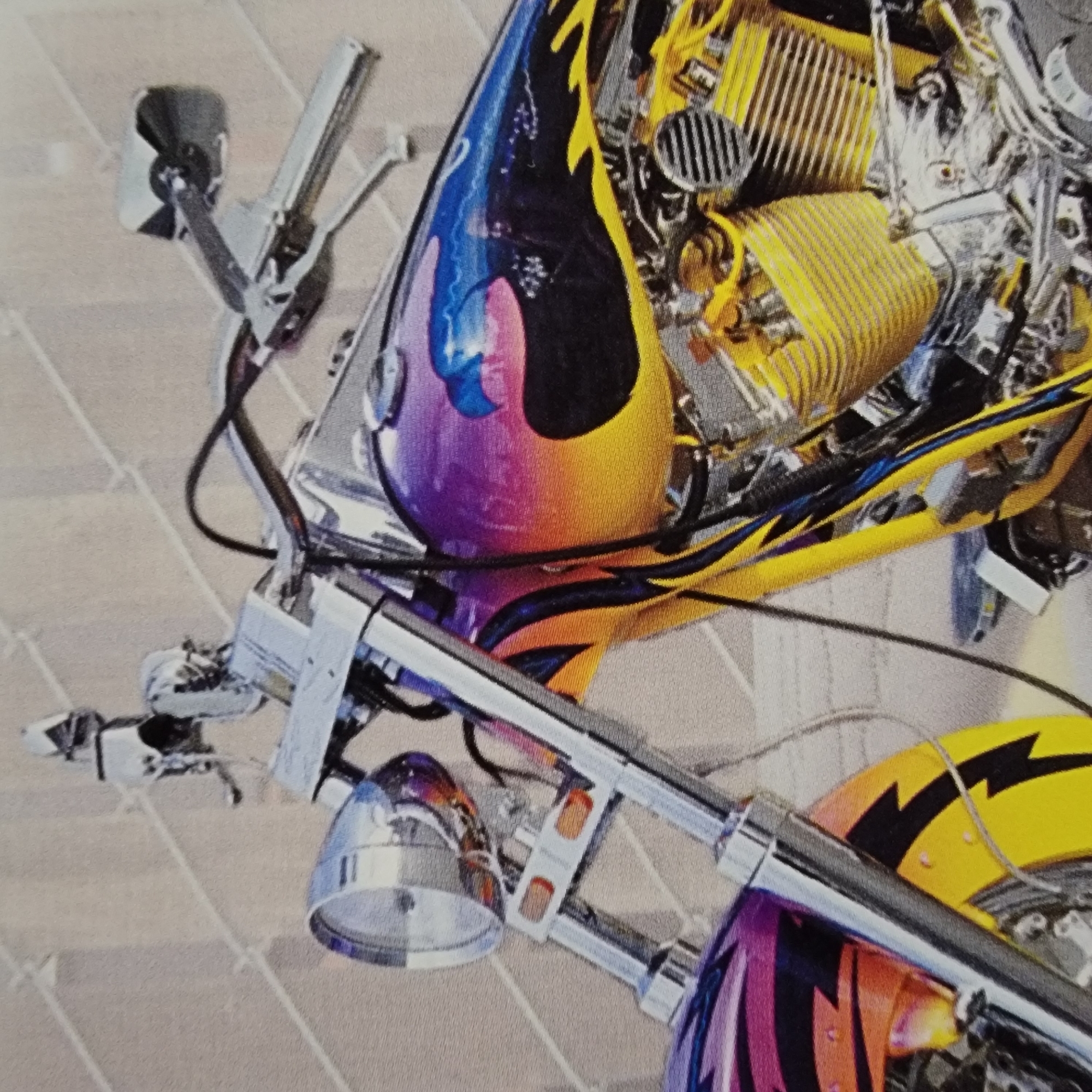 The image shows a close-up, eye-level, outdoor shot of a custom motorcycle. The motorcycle is predominantly chrome with a vibrant, multi-colored paint job featuring purple, yellow, and black flames and an eagle graphic on the fuel tank. The foreground shows the front of the motorcycle, including the headlight, front forks, and part of the front fender. The middle ground displays the engine and a portion of the fuel tank with the detailed paintwork. The background consists of a textured, light-colored wall, possibly brick or stone, with a grid-like pattern. No people or actions are visible in the image. The location is unspecified.
