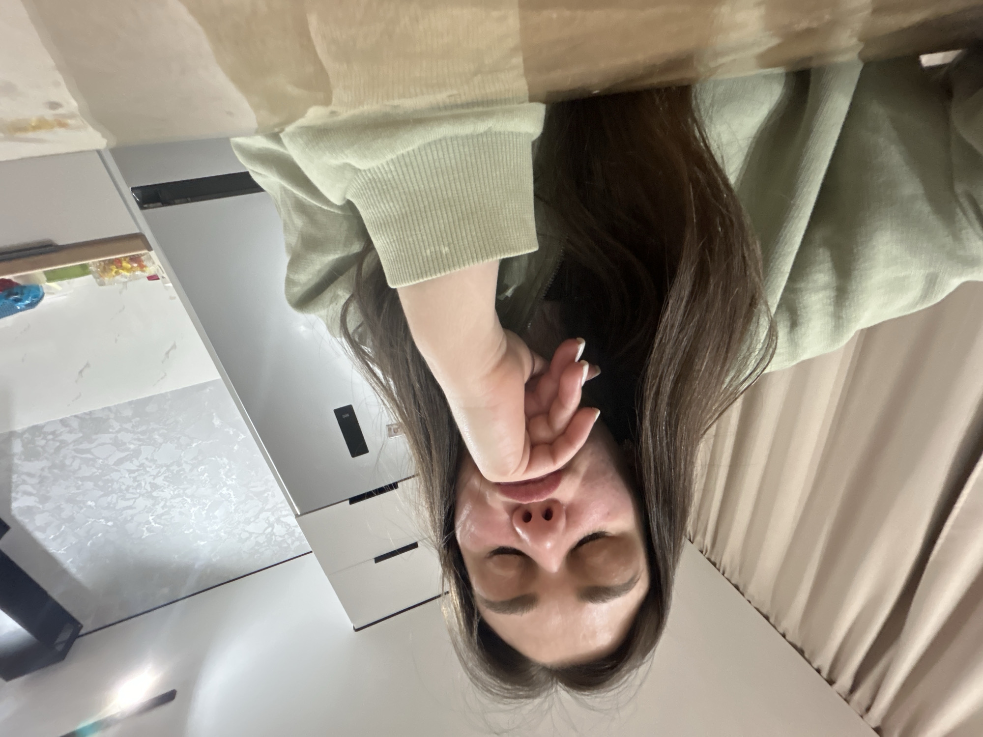 The image displays a close-up, low-angle view of a young person, possibly female, with long, dark brown hair. The image is oriented upside down, with the person's head at the bottom of the frame and their hair flowing upwards. Their eyes are closed, and their right hand, featuring nails with white tips, is covering their mouth, suggesting they might be resting, sleeping, or stifling a yawn. The person is wearing a light green or sage green long-sleeved top with ribbed cuffs. Their skin appears flushed in some areas of the face.
The background, also appearing inverted, shows a light-colored surface, possibly a table or desk, upon which the person seems to be resting their head. This surface has some light brown stains or marks. To the left of the person (when viewed upright), there are white cabinets or drawers with black horizontal handles. One cabinet reveals colorful items, possibly candies or small objects, in clear containers. Further back, a patterned white and grey surface, perhaps a wall or decorative panel, is visible. To the right of the person, a light beige or cream-colored curtain with vertical folds hangs. No visible text is present in the image. The scene is indoors and conveys a sense of quiet or rest.