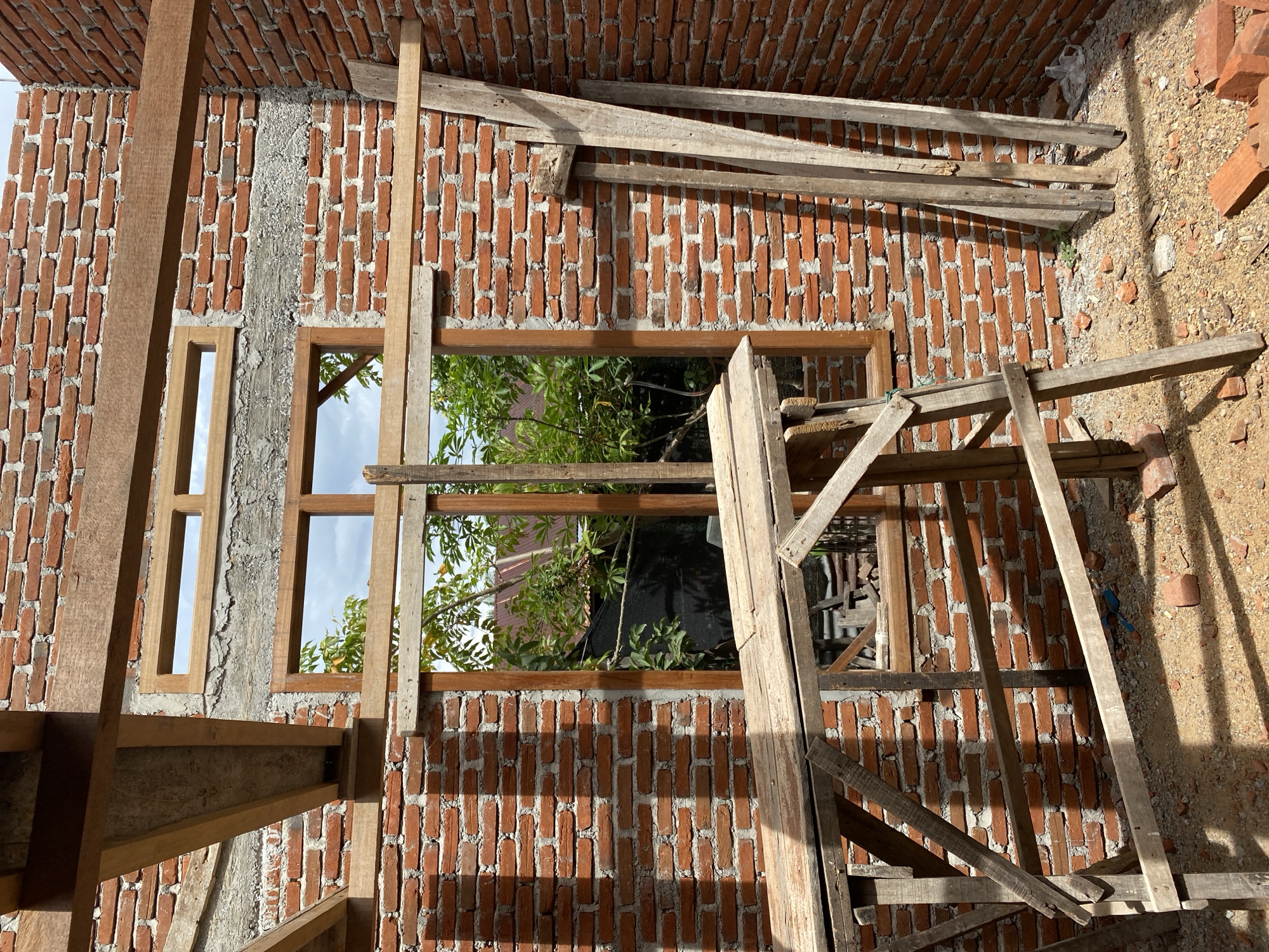 This is an exterior, eye-level, medium shot of a brick house under construction in Banda Aceh, Indonesia. The walls are made of red brick with visible mortar. A wooden window frame is installed in the wall, and above it, there is a smaller, horizontally oriented window frame. Wooden beams, likely part of the roof structure, are visible overhead. In the foreground, a wooden scaffold is erected, with planks forming a platform. Several long, wooden planks are leaning against the wall on the right side of the image. Outside the window, green foliage from trees and the roof of another building are visible. The ground is covered with dirt, gravel, and scattered bricks. The lighting suggests it is daytime, with shadows cast by the scaffold and beams.