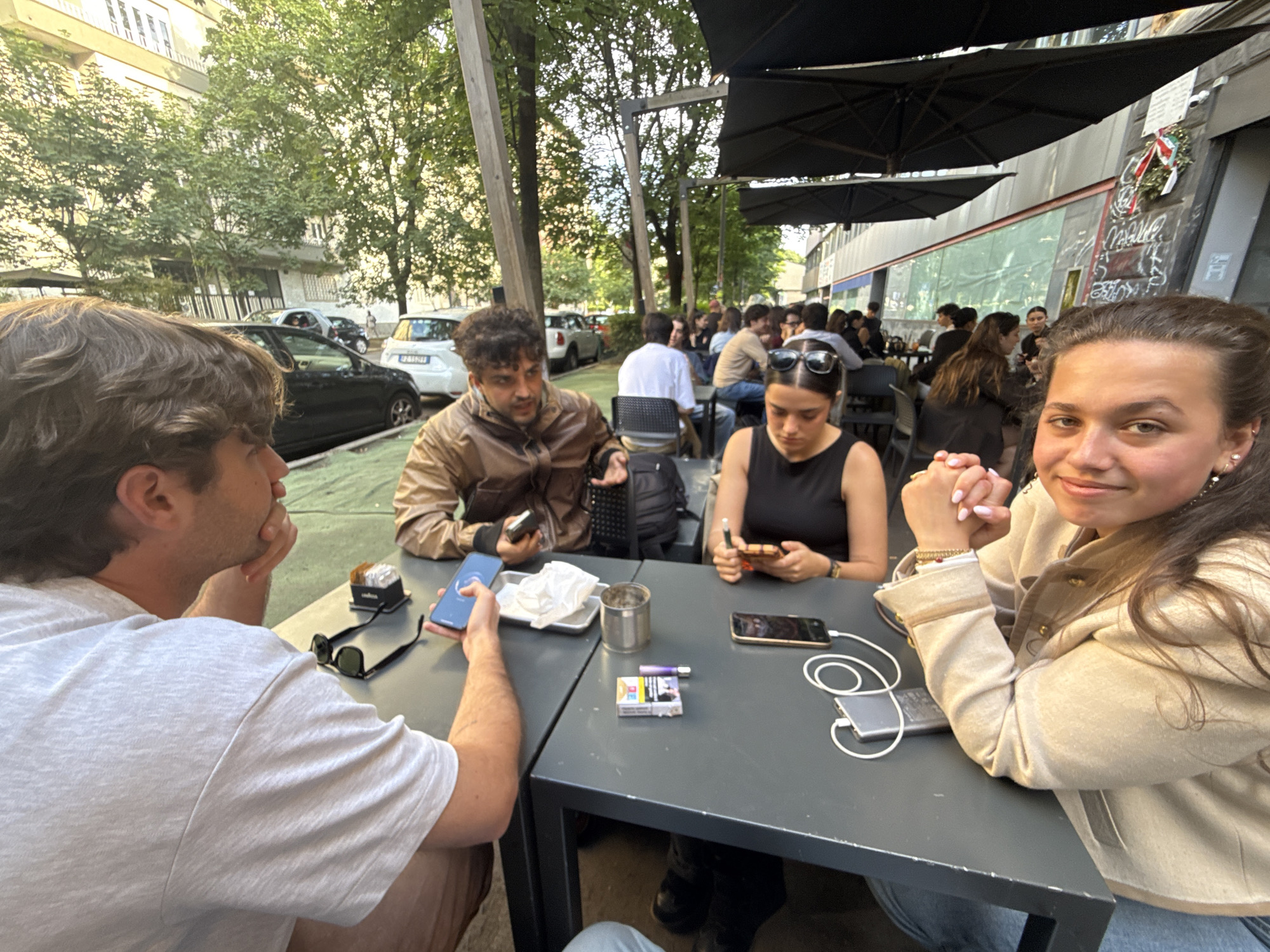 The image shows four people seated at two connected tables in an outdoor setting, likely a cafe or restaurant. The scene is situated along a street in Milan, Italy, as evidenced by the Italian flag displayed on a nearby building. The foreground features two tables with a dark gray surface.
At the first table, there are two people on the left side of the frame. A man with curly brown hair is visible mostly from the back, and he holds a blue smartphone in his right hand. Next to him are black sunglasses, a container holding napkins, a metallic can, a pack of cigarettes, and a charger.
The person directly opposite him has short brown hair and is wearing a brown leather jacket. He also is seen holding a cell phone.
At the second table, two women are seated. One woman, wearing a black top and sunglasses on her head, is looking down at a smartphone in her hand. Next to her are a portable charger and cords. The woman on the right is facing the camera and smiling, wearing a beige jacket.
In the background, additional patrons are seated at tables, creating a bustling environment typical of Milan's social scene. Trees line the street, and cars are parked nearby, adding to the urban setting. Black umbrellas provide shade over the outdoor seating area.