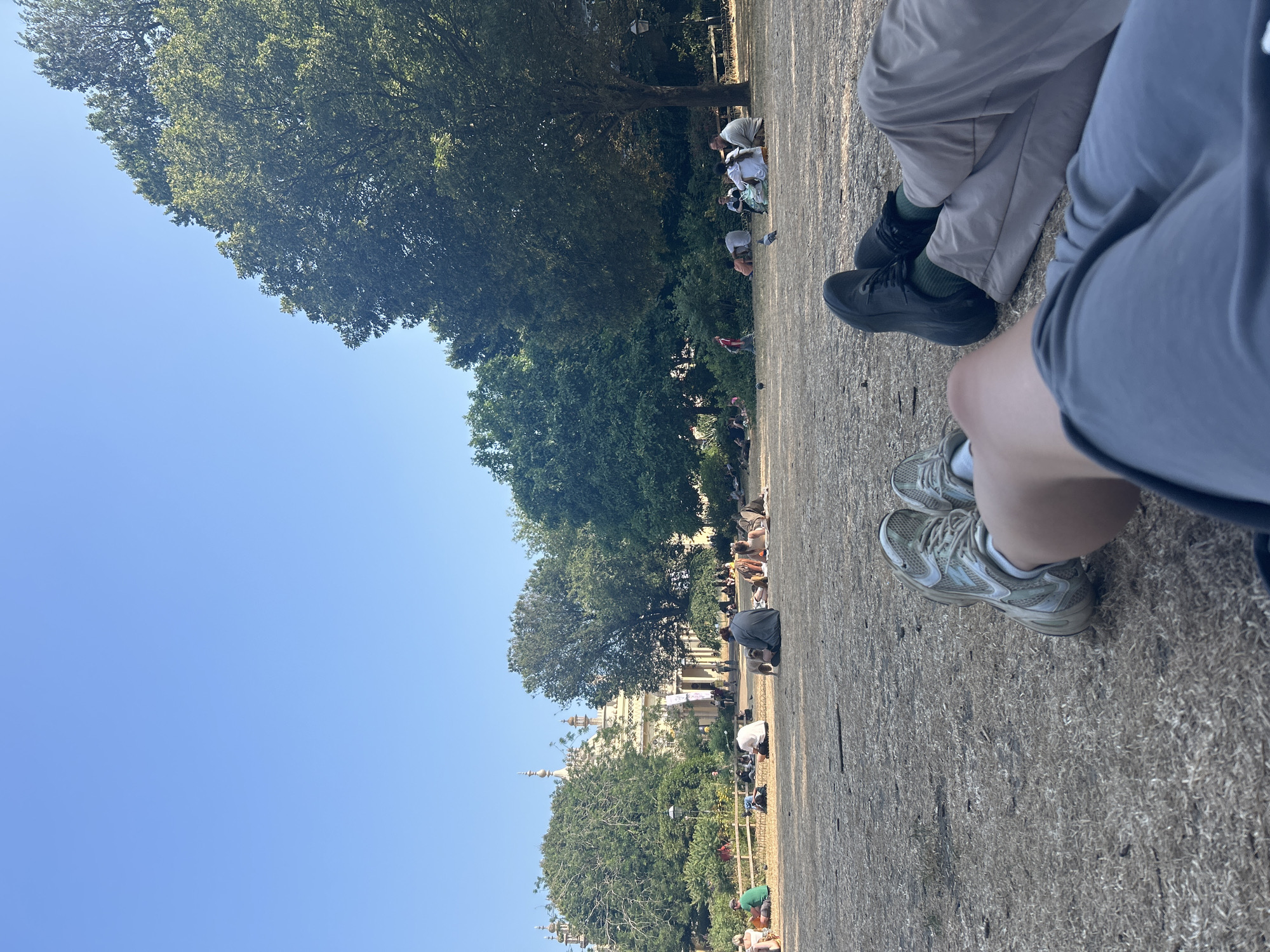 In Brighton, United Kingdom, a low-angle shot captures two individuals relaxing on dry, patchy grass in the foreground, their legs extended towards the camera. The person on the left wears light-colored athletic shoes and white socks, while the person on the right is dressed in dark sneakers, green socks, and light-colored trousers.
In the midground, a diverse group of people are scattered across the lawn, many sitting or lying down. Some are engaged in conversation, while others appear to be relaxing or picnicking. A small, light-colored dog is visible near the center of the frame, interacting with people.
Further in the background, a notable building with distinct architectural features, including domes and ornate detailing, stands amidst lush green trees. The building's light color contrasts with the surrounding foliage and the clear blue sky. The scene suggests a pleasant, sunny day in a public park, likely the Royal Pavilion Gardens given the architectural style. The overall atmosphere is relaxed and social.