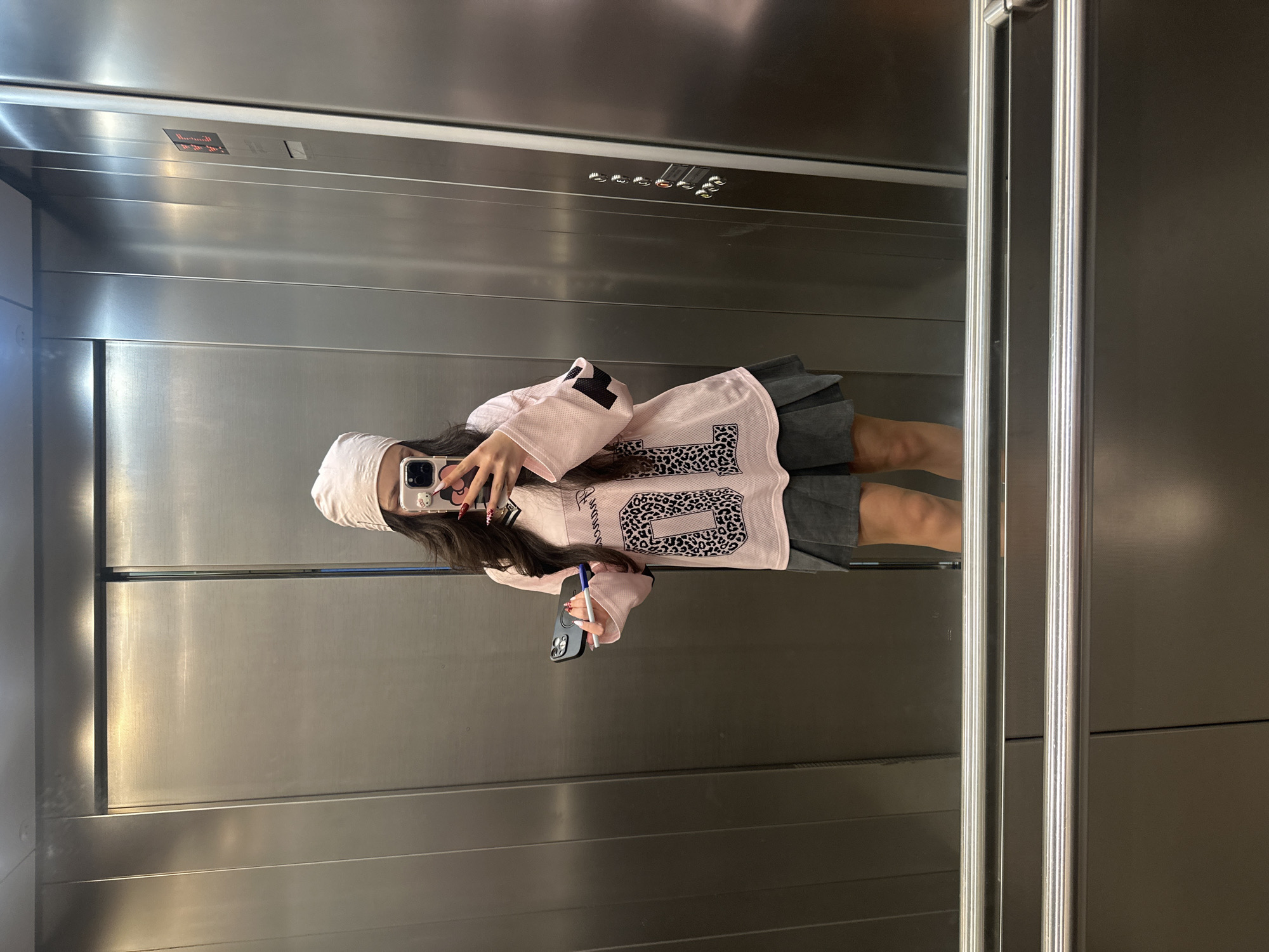 A person is reflected in a mirrored elevator in Yerevan, Armenia. The elevator is made of brushed metal with two vertical handrails on the right. The elevator's control panel with indicator lights is visible above. The person in the reflection is wearing a white headscarf, a pink long-sleeved shirt with a leopard print number "6", and a dark grey skirt. They are holding a smartphone in their right hand and a pen in their left hand. The background includes the metallic interior of the elevator.
