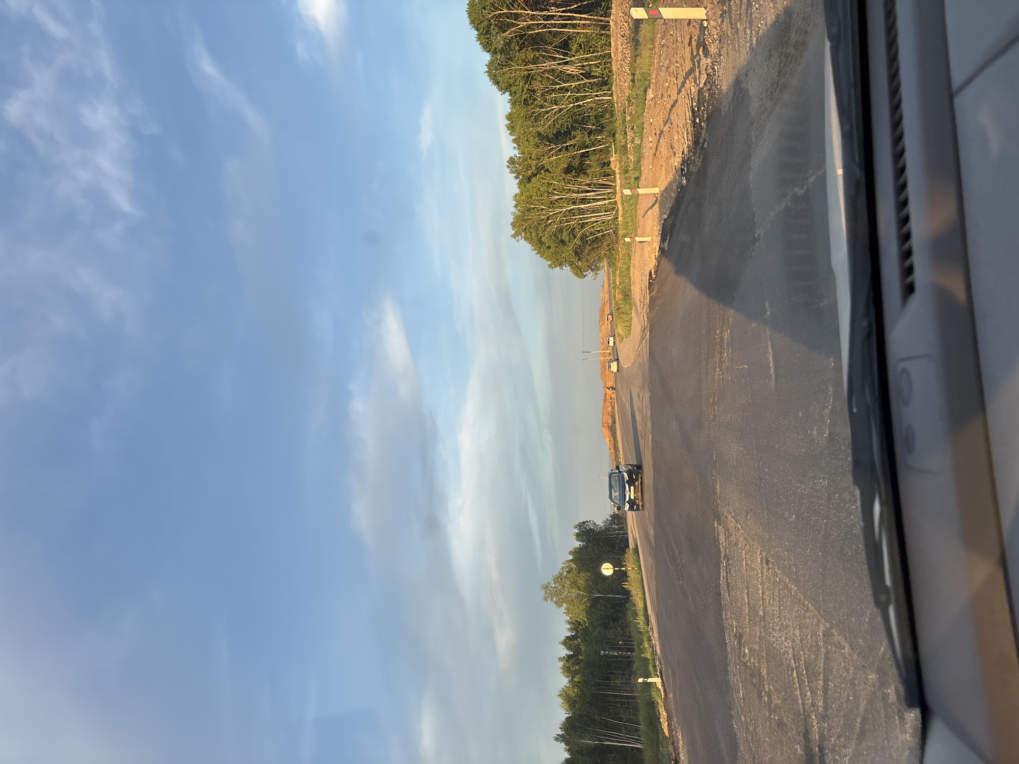 The image depicts a roadside construction scene in Plodopitomnik, Russia. A black car is visible on an asphalt road that appears to be under construction, with piles of dirt and gravel on the side. A line of trees, primarily deciduous with some evergreens, borders the construction area. The sky is a clear blue with wispy white clouds, suggesting it is daytime, likely late afternoon given the long shadows. Several road markers, white with red tops, are placed along the roadside. The perspective suggests the photo was taken from inside a vehicle, possibly looking out of a side window, with part of the vehicle's interior visible in the foreground. The overall scene conveys a sense of ongoing development and transition in a rural or semi-rural setting. No text is visible in the image.
