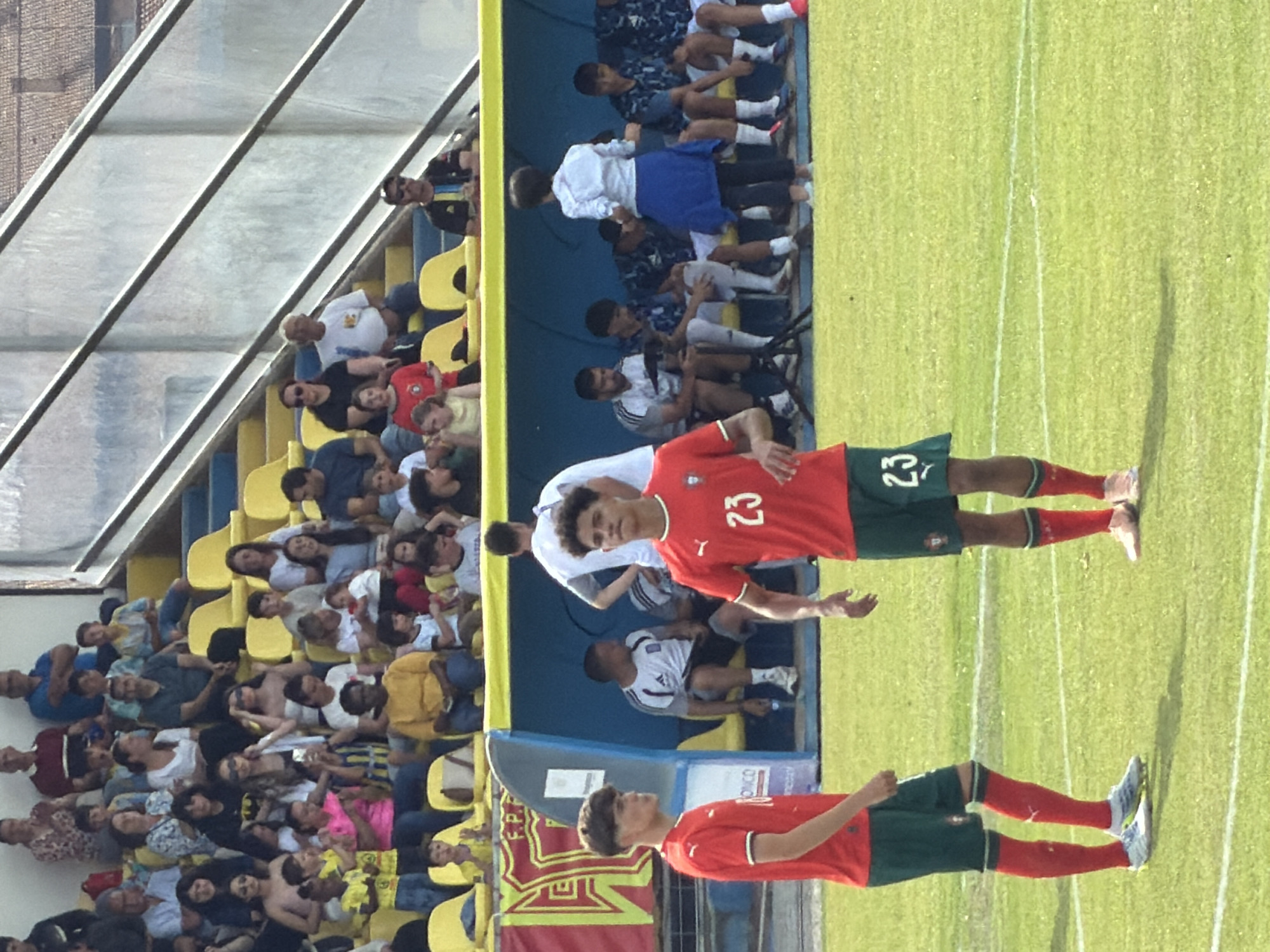 The image shows two male soccer players wearing red jerseys with the number 23 and green shorts standing on a green soccer field. The background includes a crowd of spectators sitting in yellow seats, a covered seating area, and a banner with the word "BRAGANÇA." The soccer players are standing near a sideline. The setting seems to be a soccer game or event in Bragança, Portugal.