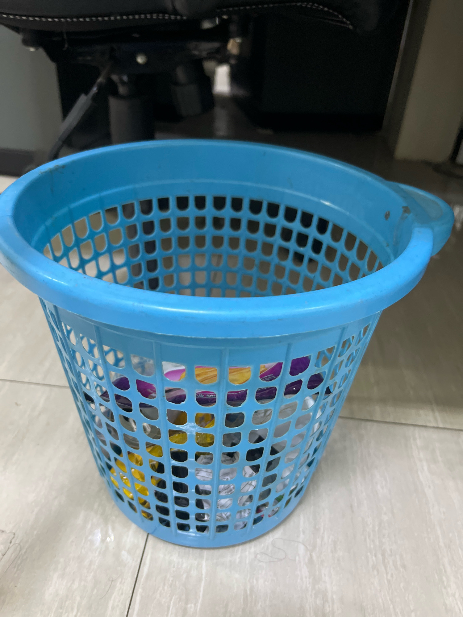A bright blue plastic waste basket with a woven pattern is the primary focus of this image. It is filled with various discarded items, including what appear to be crumpled paper wrappers and some colorful objects, possibly plastic packaging. The waste basket is positioned on a light-colored tiled floor, which shows faint lines indicating individual tiles. In the background, out of focus, is the base of a black office chair and part of a desk or cabinet. The scene appears to be indoors, likely in an office or home setting. The image was taken in Yogyakarta, Indonesia.