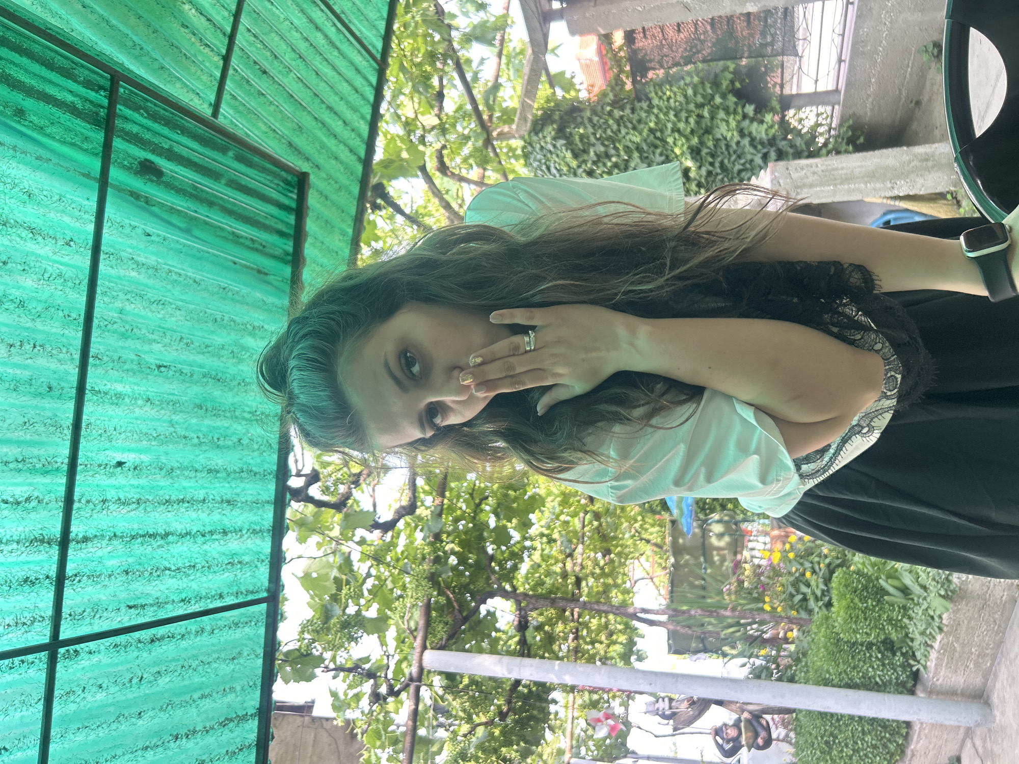 In Govedare, Bulgaria, a woman is captured from the chest up in an outdoor setting. She has long, wavy brown hair and is wearing a white top with black lace trim and a black skirt. Her left hand is raised to her mouth, covering her lips, and she is wearing a ring on her ring finger. She also has a smartwatch on her left wrist. The background reveals a lush green environment with trees and foliage, suggesting a garden or yard. Above her, a green corrugated plastic roof is visible, supported by metal beams. The lighting indicates it is daytime.