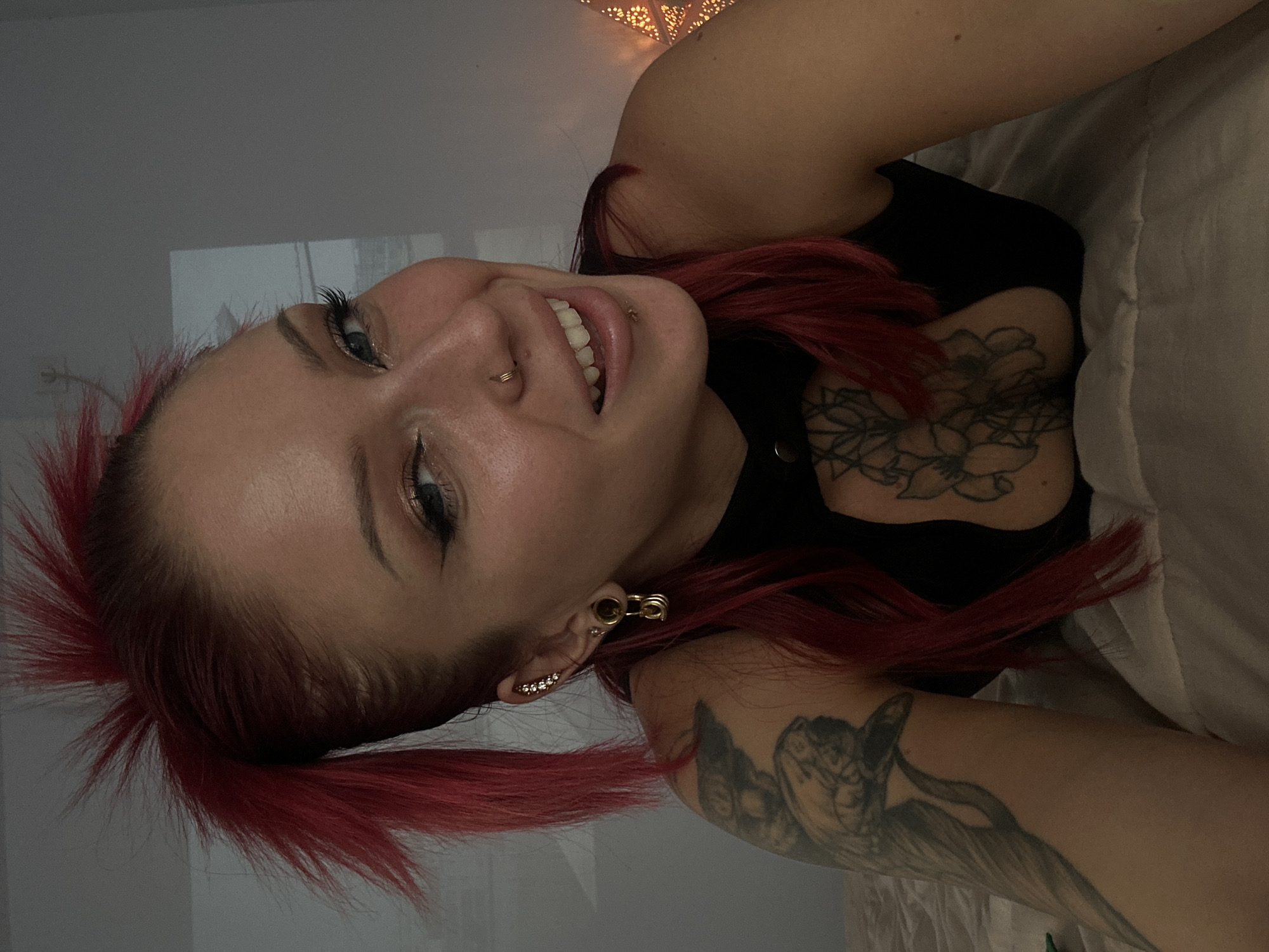 The image depicts a woman with red hair, fair skin, and multiple piercings, lying on what appears to be a bed. She is wearing a black top and has tattoos on her chest and arm. The background is blurred, suggesting an indoor setting. There is no discernible information about Dalung, Indonesia, or any indication that the image was taken there.