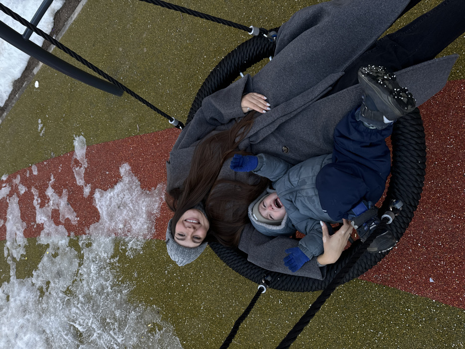An overhead photograph captures an adult female and a young child situated within a black, woven basket swing on a playground surface in Vniissok, Russia. The adult female, lying on her back, wears a grey knitted beanie, a grey long-sleeved coat, and a grey turtleneck. She has long dark hair and is looking upwards towards the camera with a wide smile. Her left hand is positioned near the child's lower back. The young child, dressed in a grey hooded winter jacket, blue gloves, and dark blue winter pants, is lying next to the adult, exhibiting a distressed facial expression with an open mouth consistent with crying or screaming, and raised arms. The playground ground cover features olive green and reddish-brown speckled rubberized material. Patches of melting snow and ice are visible on the ground, particularly in the upper left and right areas of the image. The child's dark winter boots are equipped with small white studs on the soles.