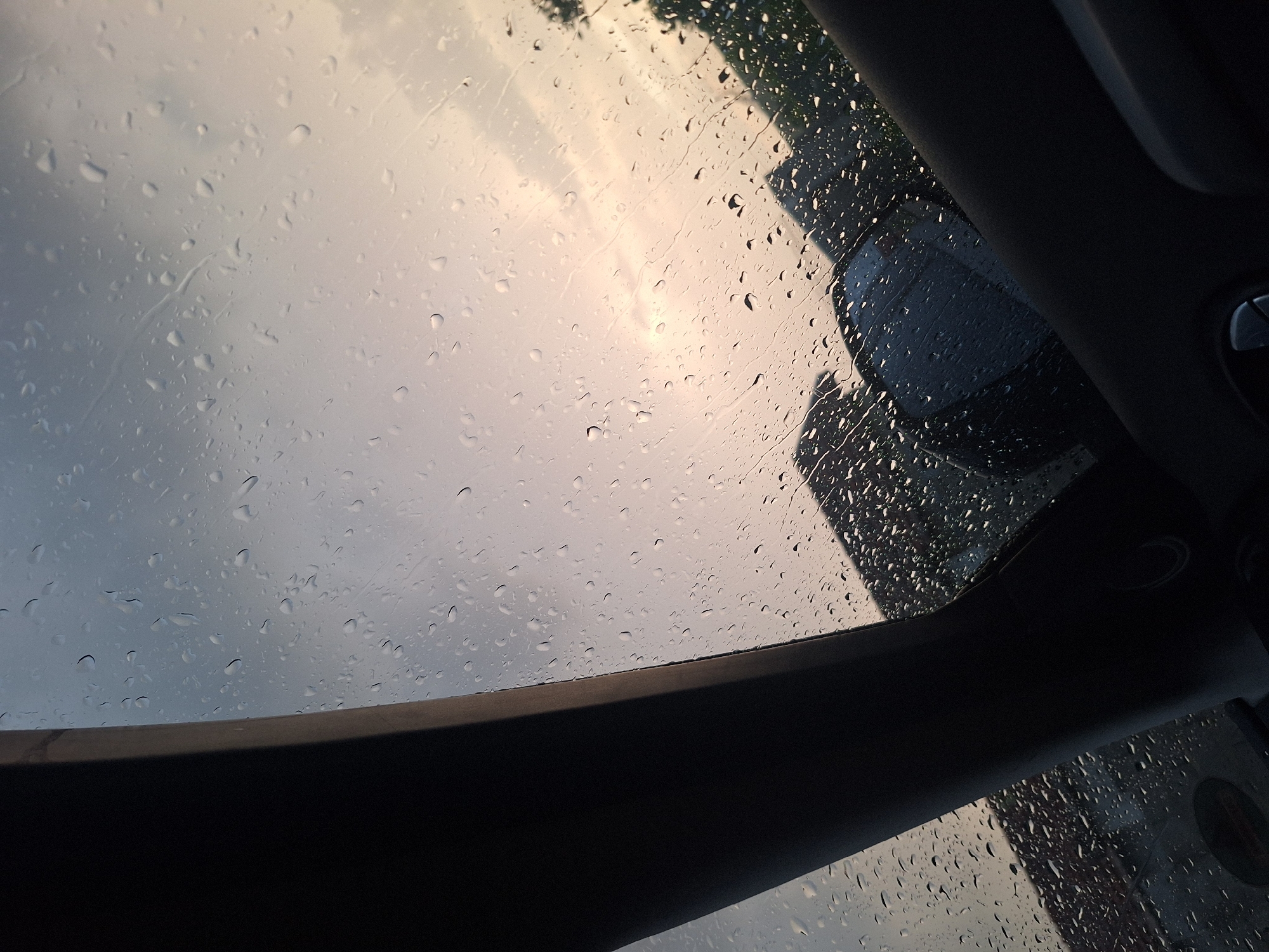 The image captures an oblique view from inside a vehicle looking through a heavily rain-covered window towards an overcast sky. Numerous water droplets of various sizes and shapes adhere to the glass surface, with some forming streaks. The sky transitions from a muted blue-grey at the top to lighter grey and warm, pale yellow-orange tones towards the bottom, suggesting either dawn, dusk, or heavily filtered light during an overcast day.
Dark, curvilinear interior components of the vehicle frame the view from the left and lower right, likely representing a structural pillar and parts of the dashboard or door panel. A circular control or button is partially visible in the lower right corner of the interior frame.
Through the rain-streaked glass, the exterior environment is visible in dark silhouettes. These include indistinct architectural structures, possibly roofs or upper sections of buildings, in the middle-left and middle-right background. Outlines of trees or dense foliage are discernible along the far right edge. A dark, indistinct shape resembling the side of another vehicle is present in the lower-middle portion of the exterior view. No individuals are visible. The location is in the city of Unknown, Unknown.