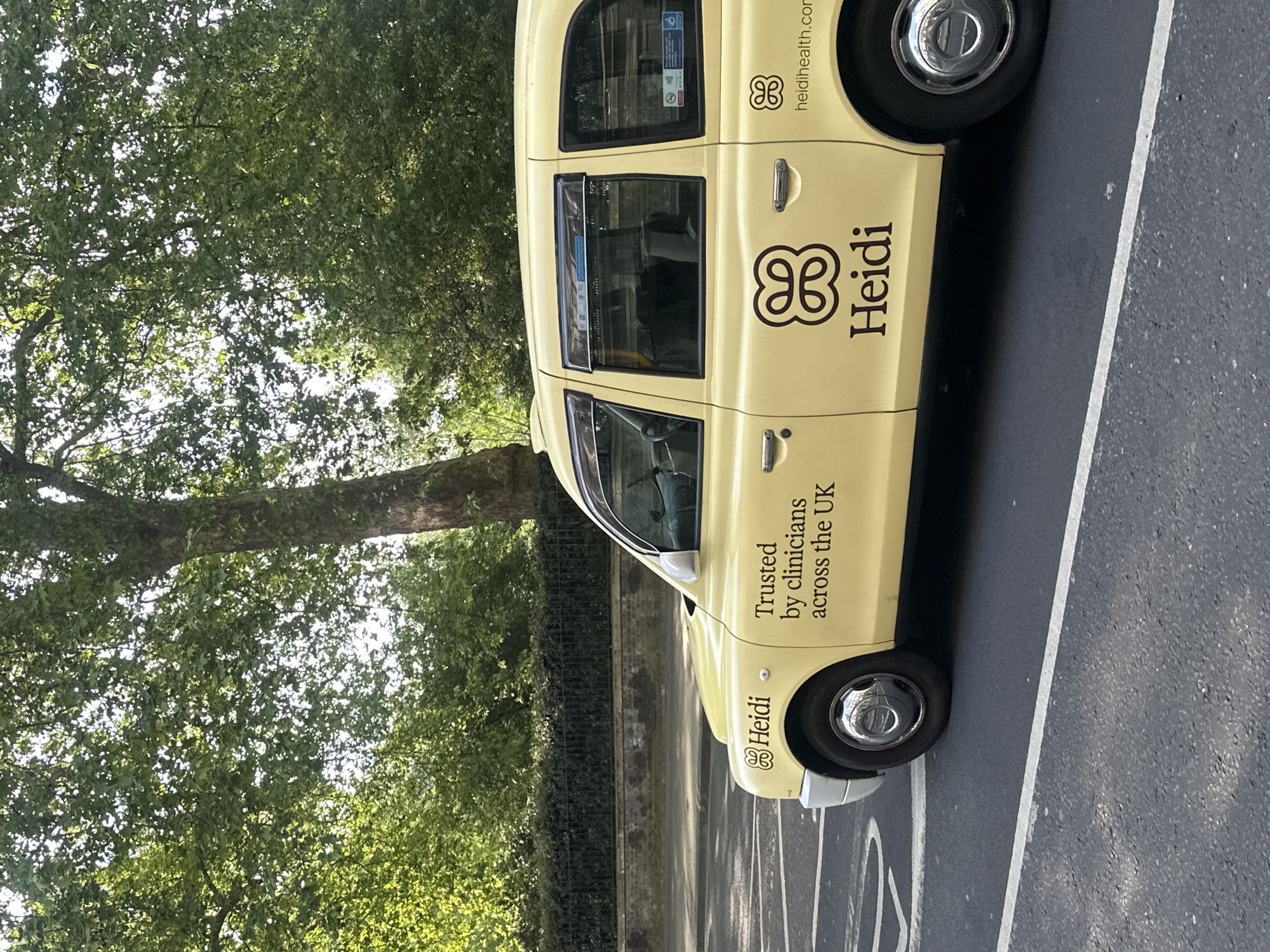 A yellow "Heidi" branded taxi is parked in a marked parking space in London. The taxi features the company's logo and tagline, "Trusted by clinicians across the UK," along with their website address. A tree and foliage are visible in the background. The setting is an urban environment in London.