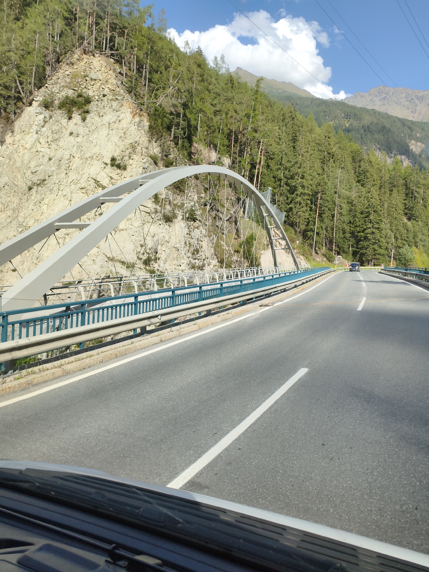 The image shows a road with a bridge in Winklen, Austria. The road is paved and has white lane markings. There is a bridge with a grey arch and blue railings that crosses over the road. On the left side of the road, there is a rocky mountain slope covered with trees. On the right side of the road, there are more trees and mountains in the background. The sky is blue with some white clouds. A dark-colored van is visible driving on the road in the distance. The photo appears to be taken from inside a vehicle, with the dashboard and windshield wiper visible in the foreground.