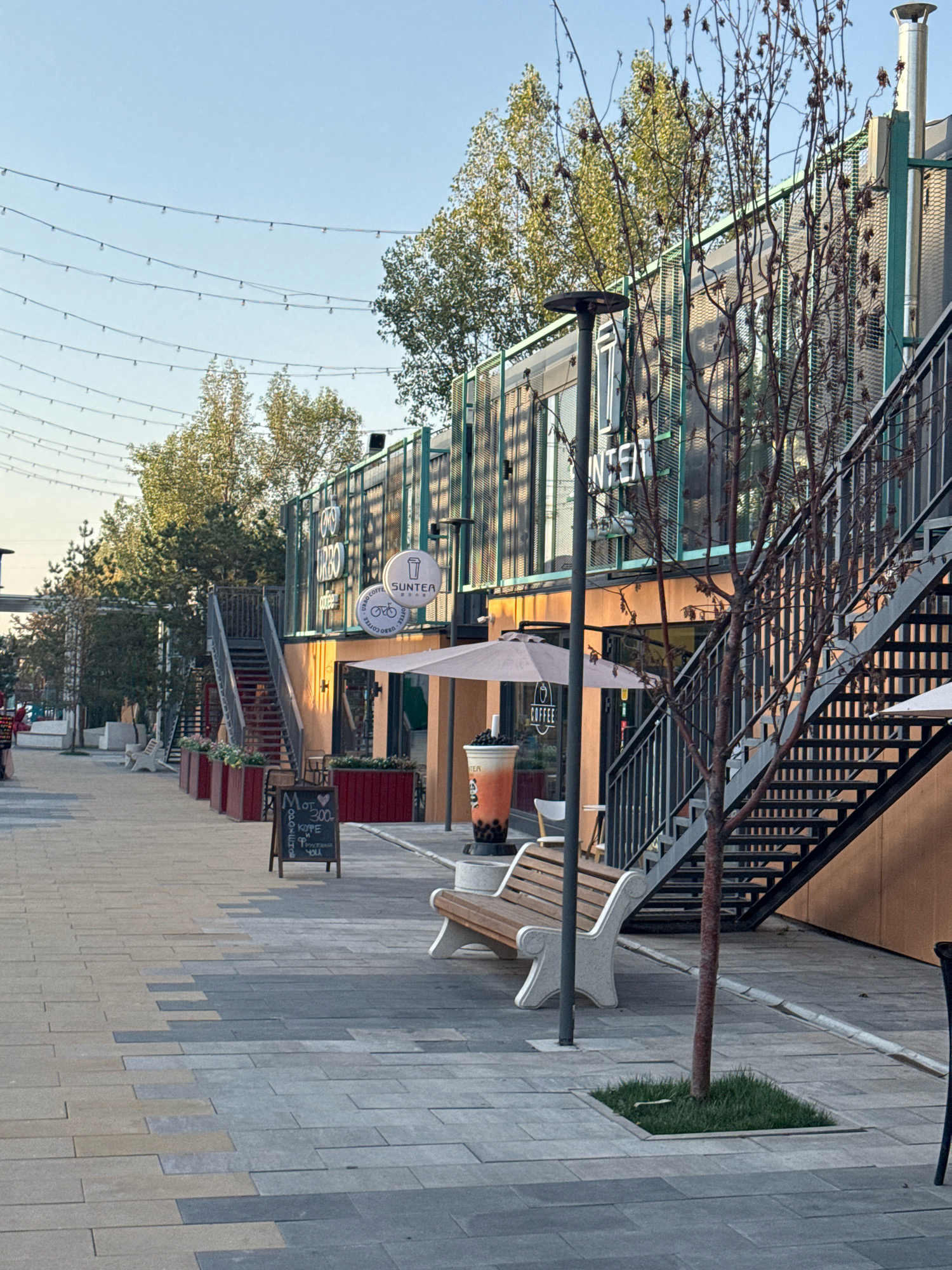 The image depicts an outdoor urban scene with a modern, multi-level structure constructed from shipping containers. The structure has an industrial aesthetic, featuring exposed metalwork and large windows. An external staircase with a black metal railing ascends the side of the building. The ground level appears to be a walkway paved with rectangular stones in varying shades of grey and beige, arranged in a geometric pattern.
To the left of the walkway, there are several planters with red fronts filled with greenery. A wooden bench with ornate white concrete armrests is placed on the paved area, facing the containers. In front of one of the ground-level units, there is a small chalkboard sign with handwritten text and a drawing of a heart. Further down the walkway, a large, clear plastic cup filled with a brown beverage and toppings, resembling bubble tea, is visible. Two light-colored patio umbrellas are deployed in front of the container units.
Overhead, strings of lights are suspended across the scene, suggesting it might be an area designed for evening gatherings. A thin tree with bare branches stands in the foreground, planted in a small square of grass. The background shows more trees with green foliage and a clear sky.
The scene is identifiable as the "Esentai Mall Container City" in Almaty, Kazakhstan, a popular spot known for its collection of cafes and shops housed in repurposed shipping containers. No people are visible in the image. The overall impression is one of a contemporary, trendy, and potentially lively urban space.