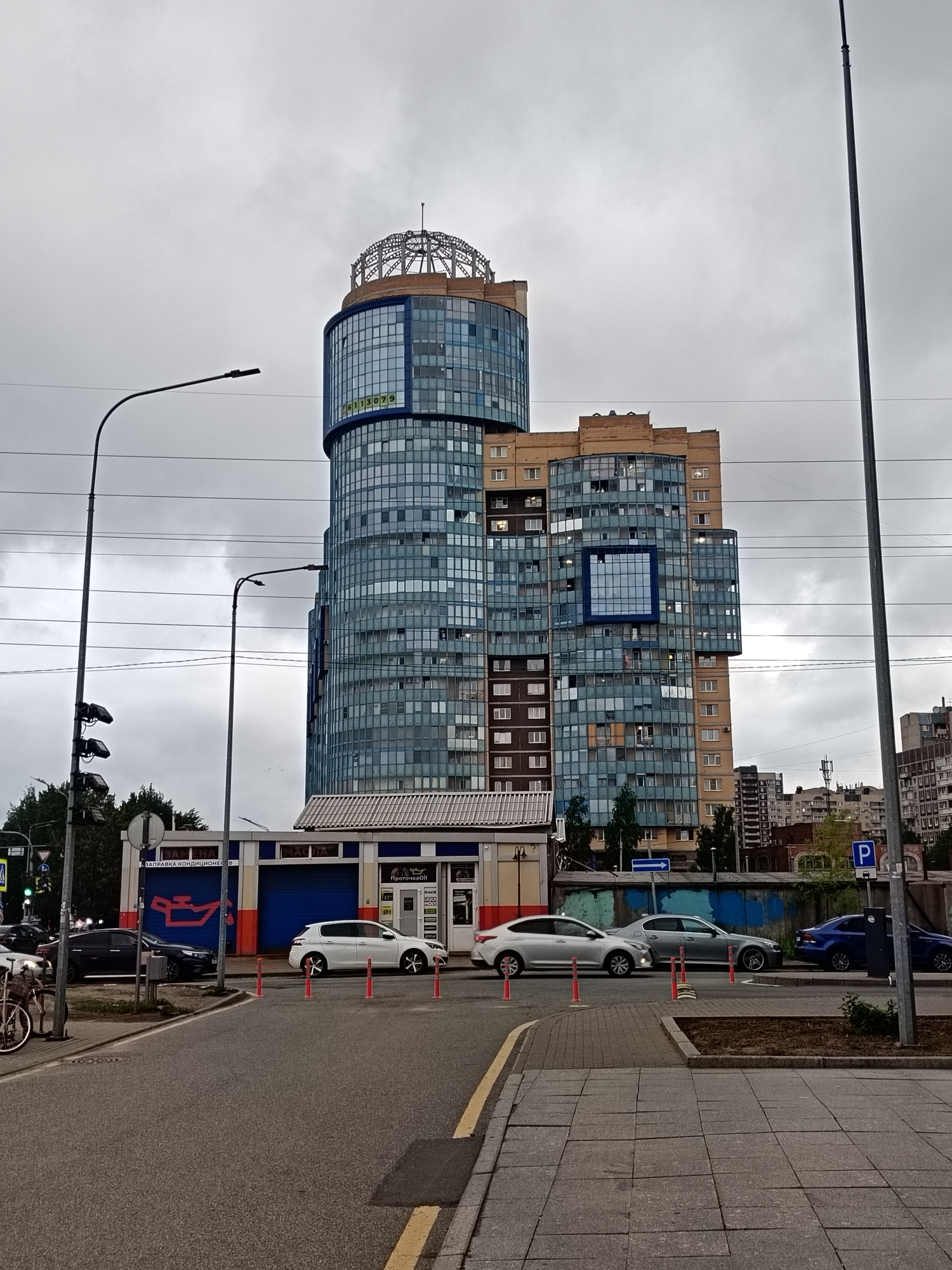 The image shows a street scene in Saint Petersburg, Russia, dominated by a tall, modern building with a cylindrical glass tower on top. The building features a mix of glass and brick elements. In the foreground, there's a street with several cars parked or driving along it. A small, low building with blue and orange accents stands next to the road, possibly a car service station. Streetlights line the road. The sky is overcast.
