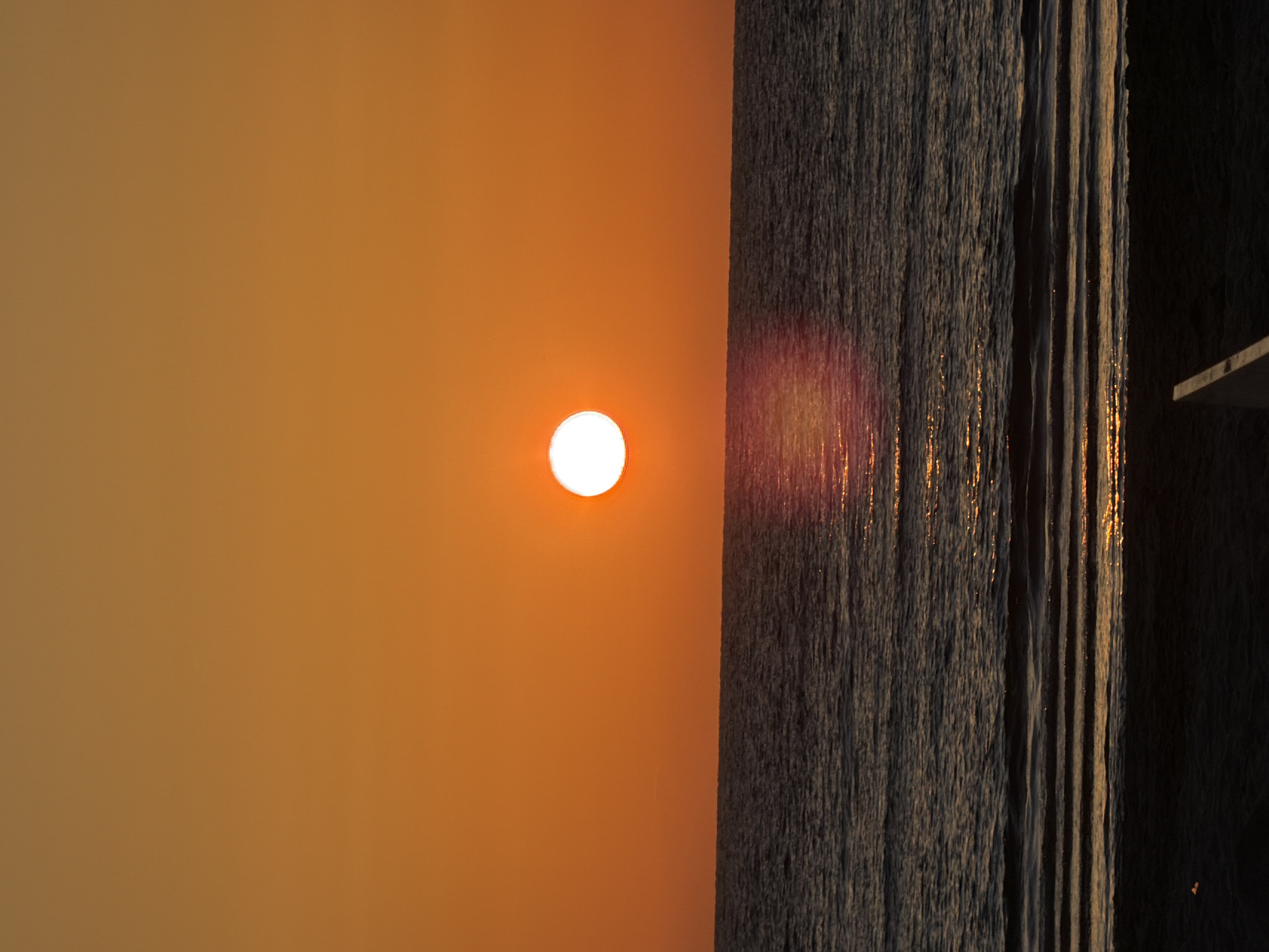 The image captures a vibrant sunset or sunrise over the sea, presenting a horizontal division between the sky and the water. The left two-thirds of the frame are filled with a clear sky, transitioning from warm, golden-orange hues near the horizon to a softer, brownish-orange further up. A brilliant, circular white sun is a central feature against this colorful sky, positioned slightly to the left of the image's vertical midline.
The right third of the image shows the surface of the sea, characterized by numerous textured ripples and small waves. Intense, elongated reflections of the sun's light streak across the water in vibrant orange and red tones, creating shimmering vertical lines. A distinct red lens flare is also visible over the water surface, adding to the warm glow. In the far right, a dark, angular structure, possibly part of a pier or a platform, is partially visible, extending into the scene and silhouetted against the water. The scene evokes a sense of calm and beauty. No text is visible in the image. Given the context, this serene seascape is likely captured in Tel-Aviv, Israel.