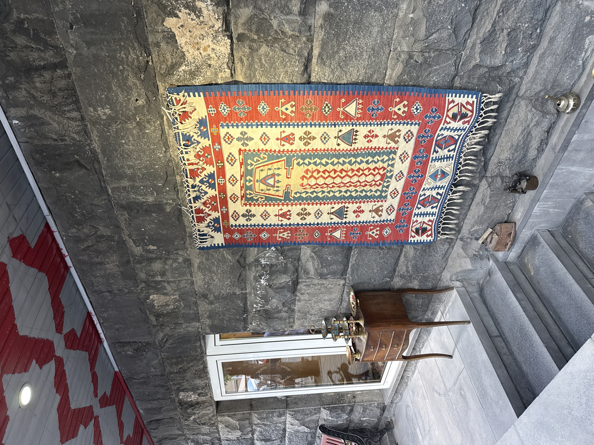 This media file captures a detailed view of an outdoor or semi-outdoor area, likely a decorated wall and surrounding space in Yerevan, Armenia. The dominant feature is a vibrantly patterned kilim or flat-weave rug, primarily red, blue, cream, and white, with intricate geometric designs and fringed edges. It is prominently displayed vertically on a dark grey, rough-hewn stone wall, which forms a significant part of the background and extends into the flooring and steps.
To the right of the rug, on the stone wall, are small metallic fixtures, including a brass-colored bell-like object and a darker, more industrial-looking piece. Below the rug, a small, ornate wooden cabinet or table with drawers stands against the stone wall, supporting a decorative metal vessel, possibly a samovar or a tea/coffee dispenser, adorned with small cups.
A modern white-framed window is set into the stone wall beneath the wooden table, reflecting elements from its surroundings, including some faint text that appears to read "ART SHOP". Stone steps lead downwards from the right side of the frame, and near the steps, a rusty old iron is visible on the stone surface. On the far left, a distinct wall with a red and white geometric pattern is visible, contrasting with the dark stone.
The scene suggests a decorative display, possibly at the entrance of a shop or a cultural establishment, emphasizing traditional craftsmanship and design. No people are visible. The natural lighting indicates it is daytime. The image is oriented sideways, with the wall acting as the vertical element and the floor/steps appearing to the right.
