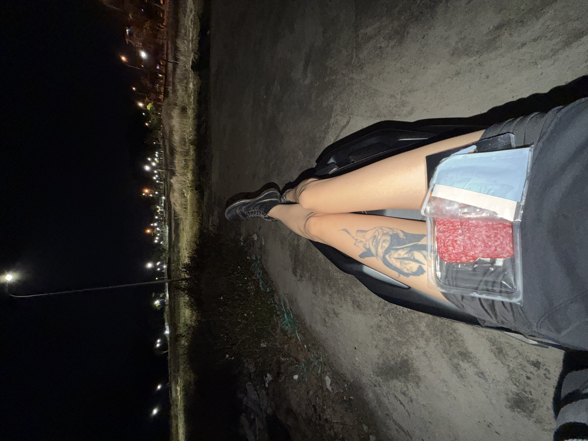 A long shot, taken outdoors at night, shows a person sitting on a black motorcycle, facing toward the upper right. The person's legs are visible, crossed at the ankles, and pointing toward the left. Both legs are exposed from the mid-thigh down. The right leg features a black tattoo on the thigh. A clear plastic food container, holding what appears to be salami, sits on the person's right thigh. The person is wearing dark clothing. The setting appears to be an unpaved shoulder adjacent to a road, with grass and debris visible. A row of lights is visible in the far background on the left. The location may be Đà Nẵng, Vietnam.