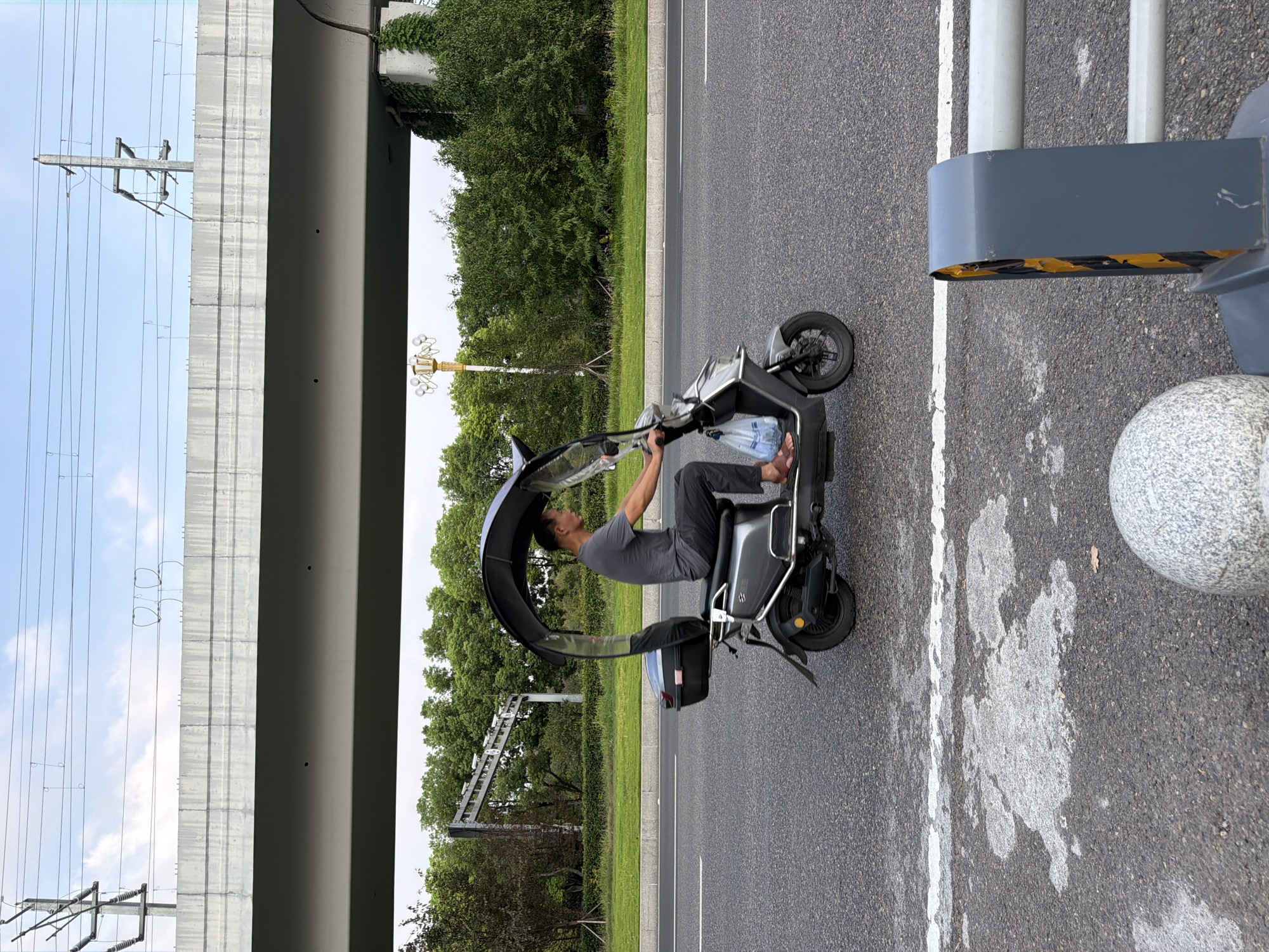 A side profile of a man riding a gray electric scooter with a canopy is captured on a paved road in Jiangjin, China. The man, wearing a dark gray t-shirt and dark pants, is seated on the scooter and appears to be holding onto the handlebars. The scooter has a black storage box mounted on the rear and a transparent windshield extending from the canopy. A plastic bag filled with blue items is attached to the front of the scooter.
The road is asphalt with a white line marking the lane. In the foreground, a section of a gray barrier with yellow hazard stripes and a speckled concrete bollard are visible. In the background, lush green trees and vegetation line the side of the road, with a large concrete overpass and overhead power lines dominating the upper portion of the image. The sky is partly cloudy with a pale blue hue. The overall scene depicts an outdoor urban environment.
