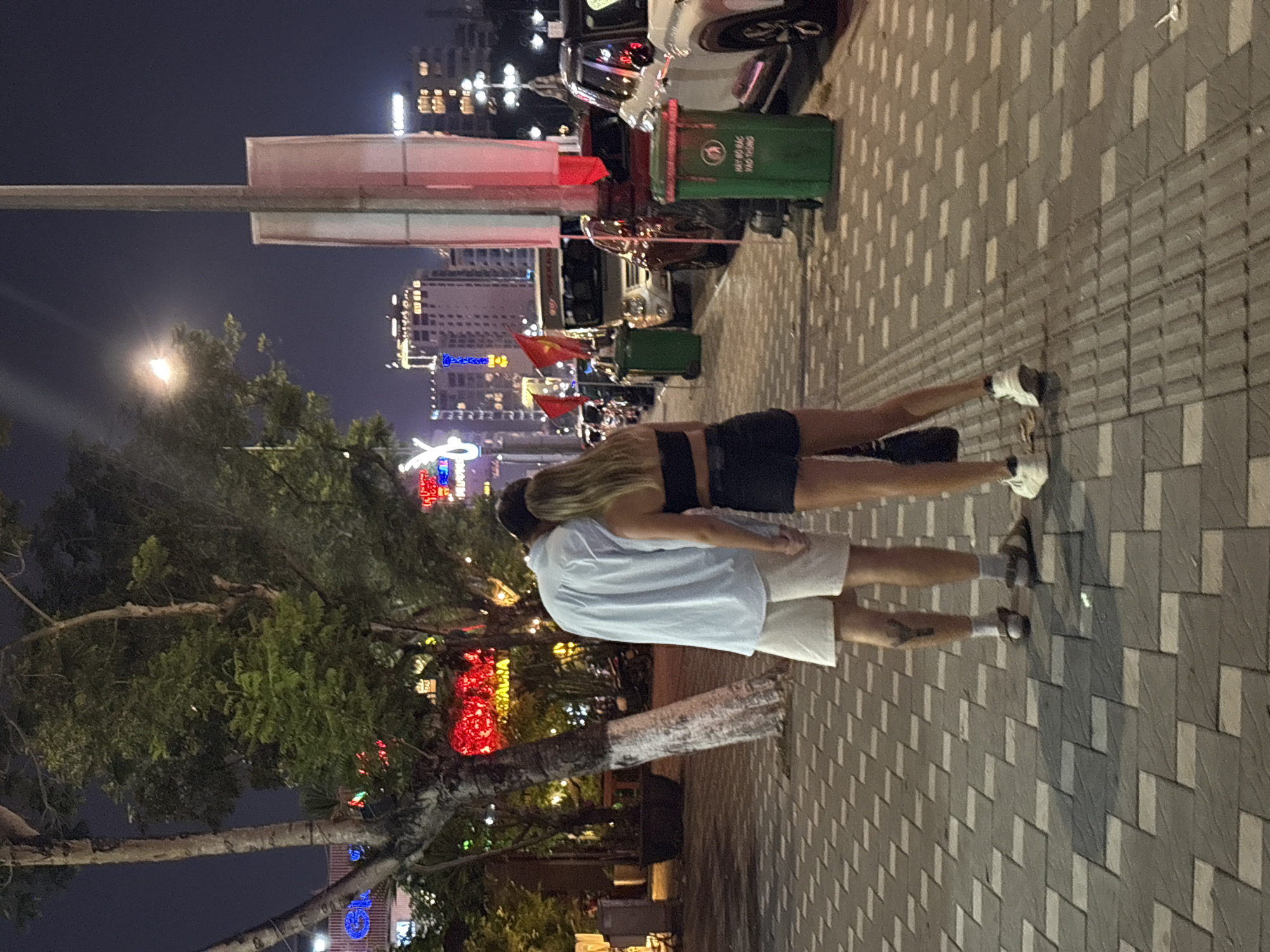 The image depicts a vibrant night scene on a city street in Đà Nẵng, Vietnam, featuring two individuals standing on a sidewalk. The perspective is slightly elevated, looking down the street. The sky is dark, indicating nighttime, with numerous artificial lights illuminating the scene.
In the foreground, two people are standing with their backs to the camera. One person, on the right, has long blonde hair and is wearing a black crop top and black shorts, paired with white sneakers. The other person, on the left, wears a light blue short-sleeved shirt, light-colored shorts, and sandals. Their arms appear to be linked or held, suggesting they are a couple or close companions.
The sidewalk beneath them is composed of a patterned block paving in shades of grey and light grey. To their left, a large tree with green foliage and a trunk partially painted white is visible, adorned with small decorative lights. In the background, beyond the sidewalk and a row of parked motorbikes and a car, tall city buildings rise, their windows glowing with interior lights and some featuring illuminated signs.
Notable details confirming the location include several red Vietnamese flags with yellow stars, seen hanging from poles or structures in the mid-ground. Also, a prominent green trash bin features the Vietnamese text "HÃY BỎ RÁC VÀO THÙNG," which translates to "Please put trash in the bin." The scene conveys a relaxed evening atmosphere in a bustling urban environment.