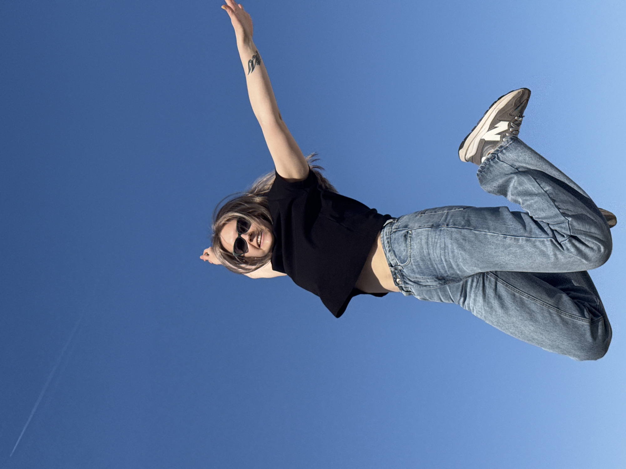 A woman wearing a black t-shirt, blue jeans, and gray sneakers with white accents is seen from a low angle against a clear blue sky. She is wearing sunglasses and has her arms outstretched. She has a tattoo on her left forearm. A faint contrail is visible in the lower left portion of the sky. The location is unknown and there is no information given on the city.