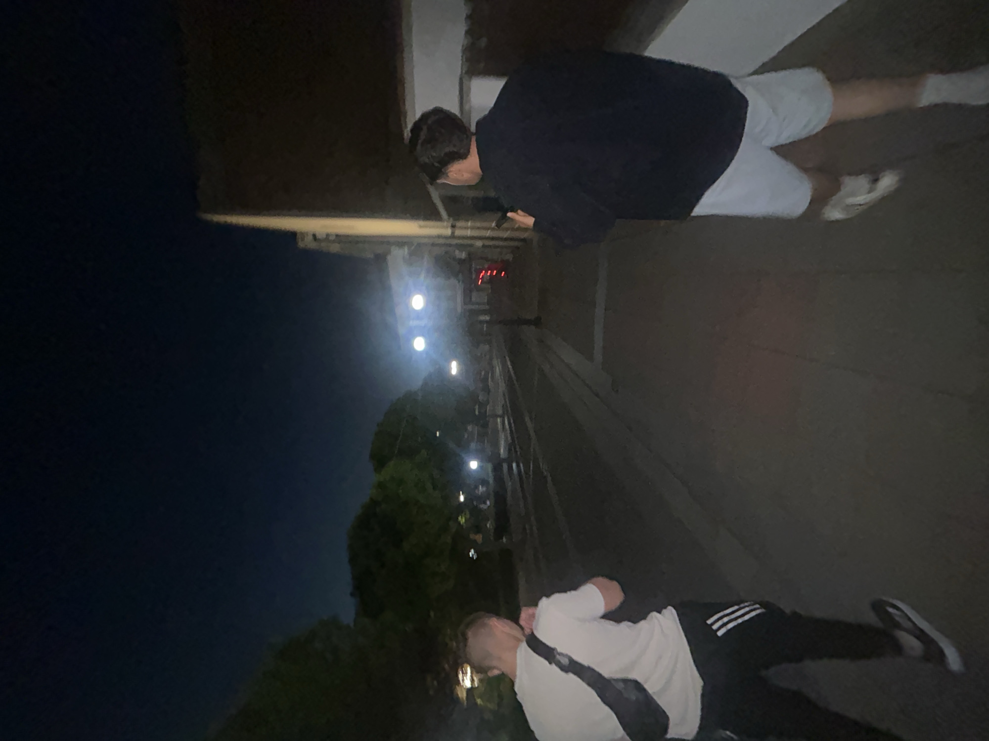Two people are visible in an outdoor setting, likely a street or sidewalk at night. One individual is wearing a dark top and light shorts, appearing to look at a phone. The second person is wearing a white shirt and black pants with white stripes and has a bag slung over their shoulder. There are streetlights and trees in the background. The exact location in Elhovo, Bulgaria, is not discernible from the image.