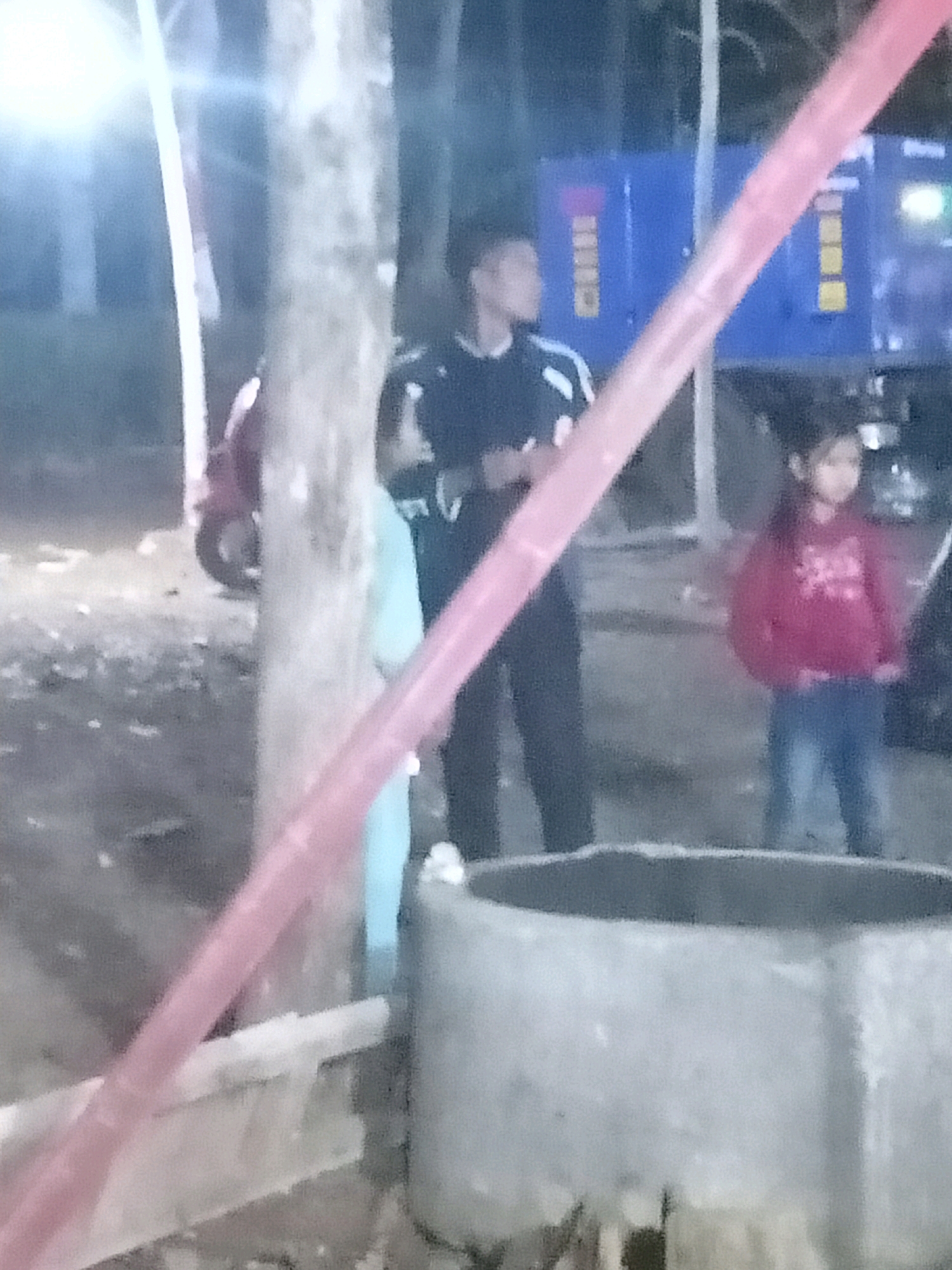 The image captures a dimly lit outdoor scene, possibly at night, with a group of people present. In the foreground, a large cylindrical concrete structure, potentially a well or a planter, is visible. A pink plastic pole or barrier diagonally crosses the frame from the left foreground towards the upper right.
Behind the concrete structure and the pole, several individuals are discernible. A young girl wearing a red hooded sweatshirt and blue jeans stands to the right, facing slightly away from the camera. To her left and further back, a young man in a dark jacket with white stripes on the sleeves is looking to the left. Partially obscured by a tree trunk in the center, a person in a light blue garment is also present. In the background, a large blue industrial generator with yellow panels is situated. The environment appears to be an outdoor area with trees and uneven ground, suggesting a possibly rural or semi-rural setting within Tasikhargo, Indonesia. The lighting is dim and appears to be from artificial sources.
