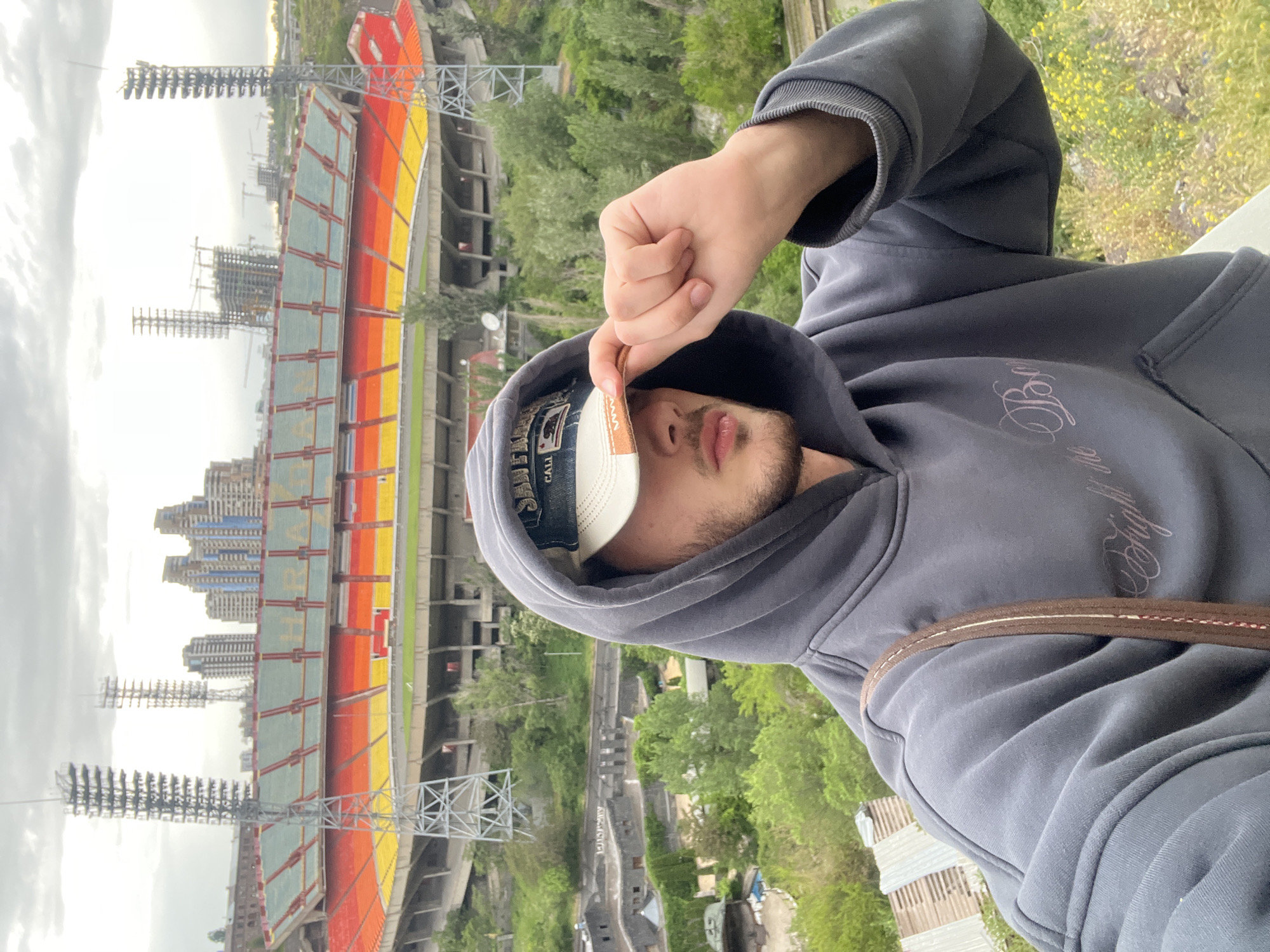 A vertical eye-level medium shot shows a person wearing a hooded sweatshirt and a baseball cap standing in Yerevan, Armenia. The person is holding a card or ticket up to their face, obscuring their eyes. Their lips and beard are visible, and their skin appears fair. The hooded sweatshirt is gray and has cursive writing on the right upper chest. The baseball cap has a denim brim and white front, with "CALI" printed on it. A brown strap crosses their shoulder.
Behind the person is a view of the city and a large stadium. The stadium is mostly red and yellow and is under construction. Tall light towers are attached to the stadium. Further in the background, numerous trees and buildings are visible, extending into the distance under an overcast sky.