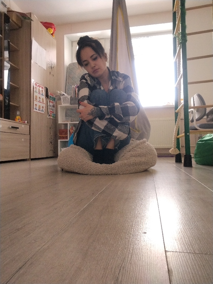 An individual is depicted seated on a light-colored, fluffy cushion on a light wood laminate floor inside a room in the city of Noviy, Russia. The person, appearing to be an adult female with dark hair styled in a bun, is wearing a black and white plaid long-sleeved shirt, dark blue jeans, and black socks. Her knees are drawn to her chest, and her arms are wrapped around them; visible tattoos are present on her left forearm. Her head is tilted slightly, and her gaze is directed forward-left.
The room is brightly illuminated by natural light entering through a window located behind the seated individual. To the left of the person is a light brown wooden wardrobe or cabinet, adorned with a red toy and several papers or charts. A small white shelving unit is partially visible behind her left side. To the right, a green and light-colored wooden climbing structure with an attached rope stands near the window. Below the window, a white radiator is present. A light fabric canopy or tent structure hangs from the ceiling near the window, and a green beanbag rests on the floor next to the climbing structure.