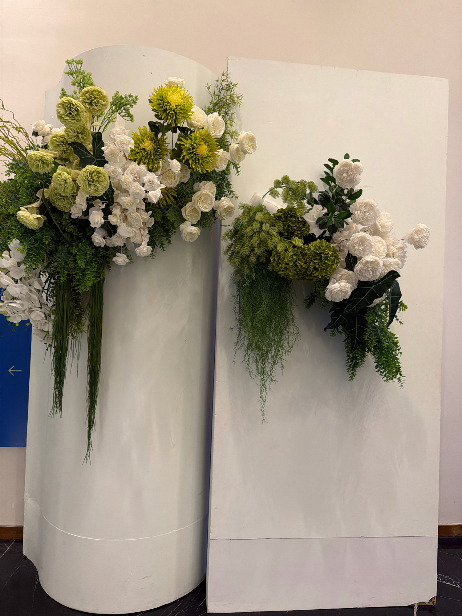 This is a photograph of two tall, pale cylindrical structures with floral arrangements on them. The structure on the left is a curved, free-standing column, while the structure on the right is a rectangular panel leaning against it. Both structures are decorated with lush floral arrangements. The arrangement on the left is more extensive, cascading down the side of the cylindrical structure with a mix of white and green flowers, including what appear to be roses, carnations, and spiky green foliage. The arrangement on the right is smaller and also features white and green flowers, with hanging vines and large, dark green leaves. The background is a plain, light-colored wall. The floor is a dark, patterned material. A blue sign with a white arrow is visible on the left side of the image, partially obscured by the cylindrical structure. The lighting is soft and diffused, suggesting an indoor setting, possibly a lobby or event space. The scene appears to be a decorative display, likely for an event or as part of interior design. The photograph does not contain any people. The city of Nha Trang, Vietnam is not visually discernible in this image.