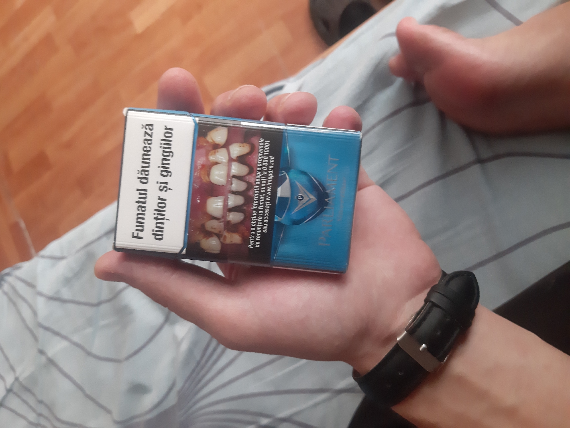 A person's right hand holds a pack of Parliament cigarettes. The cigarette pack has a warning label with an image of unhealthy teeth and gums, and text in Romanian that translates to "Smoking harms teeth and gums." The hand is wearing a black watch with a silver buckle. The background shows a striped fabric and a wooden floor. The setting appears to be indoors, possibly a home environment in Cricova, Moldova, based on the text on the cigarette pack which is in Romanian and mentions an Moldovan website.