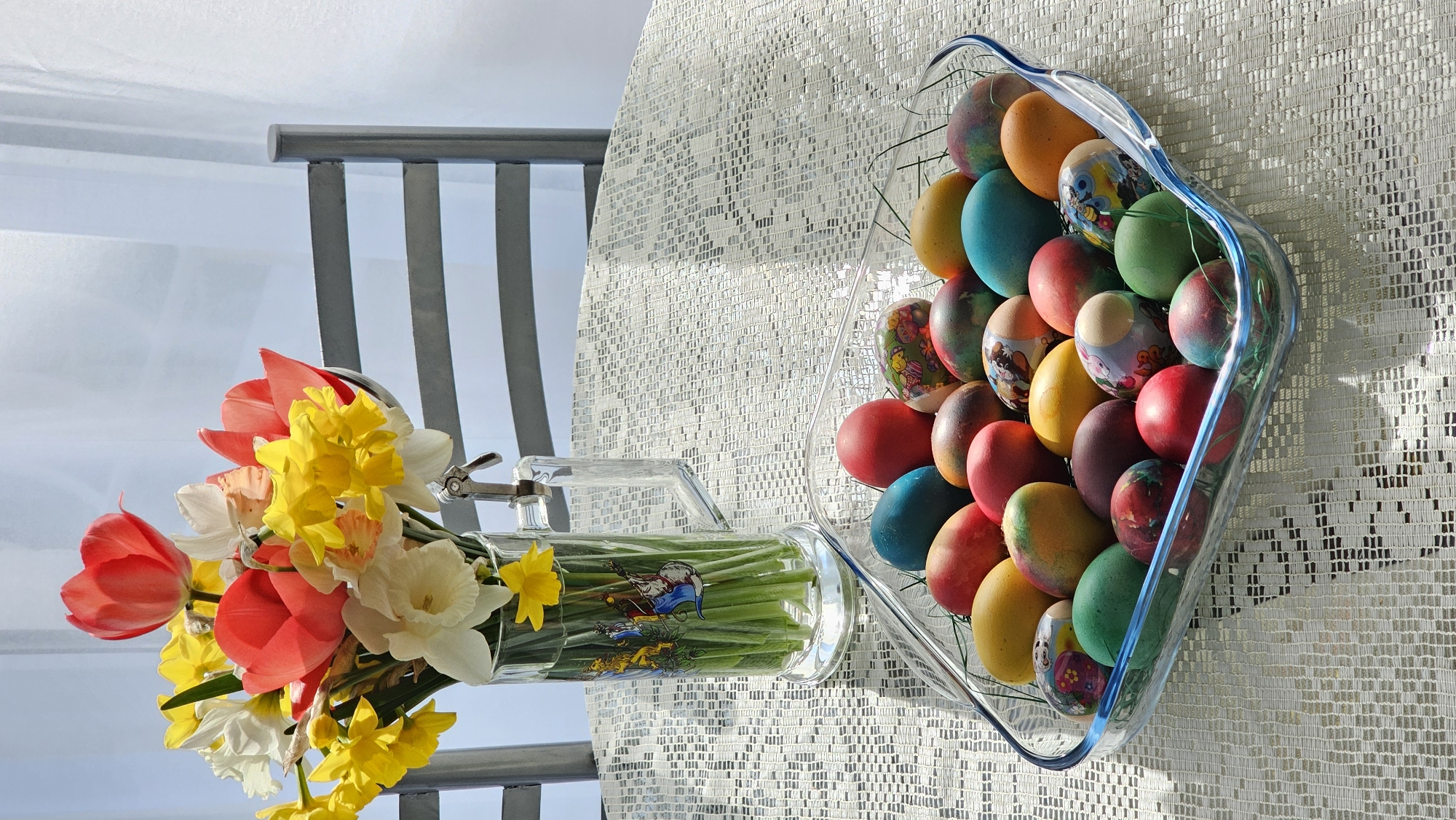 The scene depicts a tabletop arrangement. On a white, textured tablecloth sits a clear glass bowl filled with a multitude of brightly colored Easter eggs. To the left, a glass mug with a handle holds a bouquet of spring flowers including red tulips, white and yellow daffodils, and green stems. The background shows the slats of a chair and a blurred outdoor view, suggesting the setup is on a balcony or near a window. The clear light and shadows indicate a sunny day. There is no indication of location, nor information about people, subjects, actions or interactions.