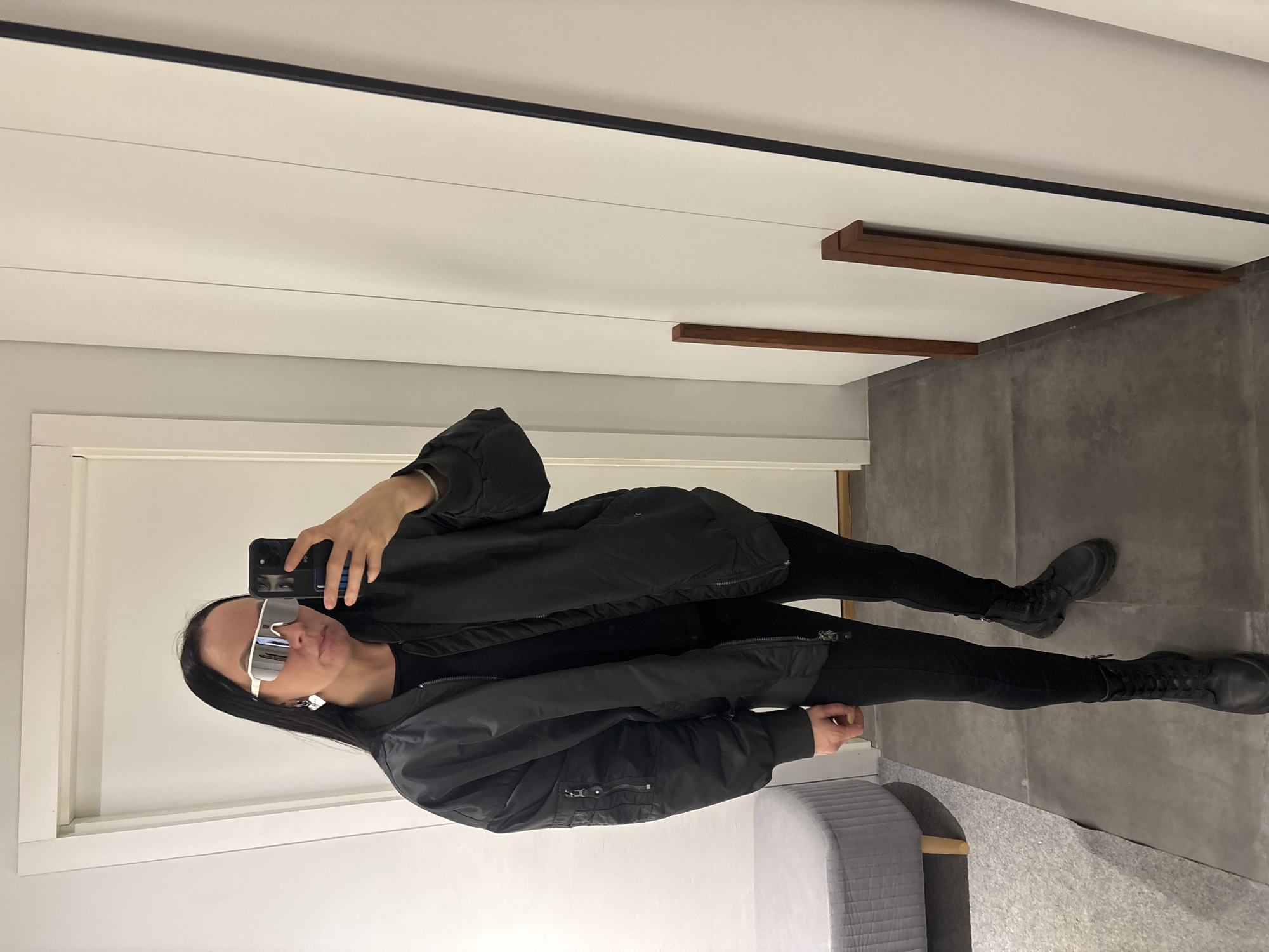 An indoor scene in the city of Chaikovsky, Russia, captures a female-presenting individual taking a mirror selfie. The subject, with long, dark hair, wears white mirrored sunglasses, silver heart-shaped hoop earrings, a black bomber jacket, black shirt, black leggings, and black lace-up combat boots. She holds a black smartphone in her right hand, angled towards a large, rectangular mirror with a thin black frame. The environment includes a dark grey tiled floor, with a light grey upholstered ottoman on a light grey carpeted area to the left. White walls are present, featuring a white door frame on the left and two vertical reddish-brown wooden panels affixed to a white surface adjacent to the mirror on the right.