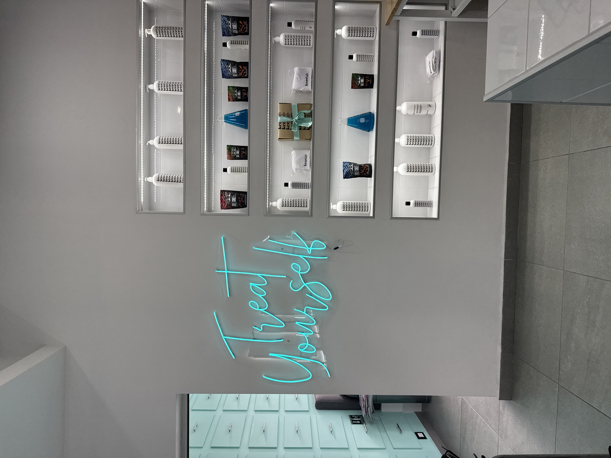 The image depicts a display of products in what appears to be a retail or salon setting in San Bernardino Tlaxcalancingo, Mexico. The main elements include a white wall with several illuminated, recessed shelving units containing various products, primarily white bottles with black text, packages of coffee labeled "XILE CHILE," and scientific-looking glass beakers. A blue neon sign that reads "Treat Yourself" is on the wall beneath the shelves. Below the sign is a blue-green door with silver handles, and a partial view of a gray chair or sofa. The floor tiles are gray. A portion of what looks like a kitchen counter or similar fixture is visible to the right.