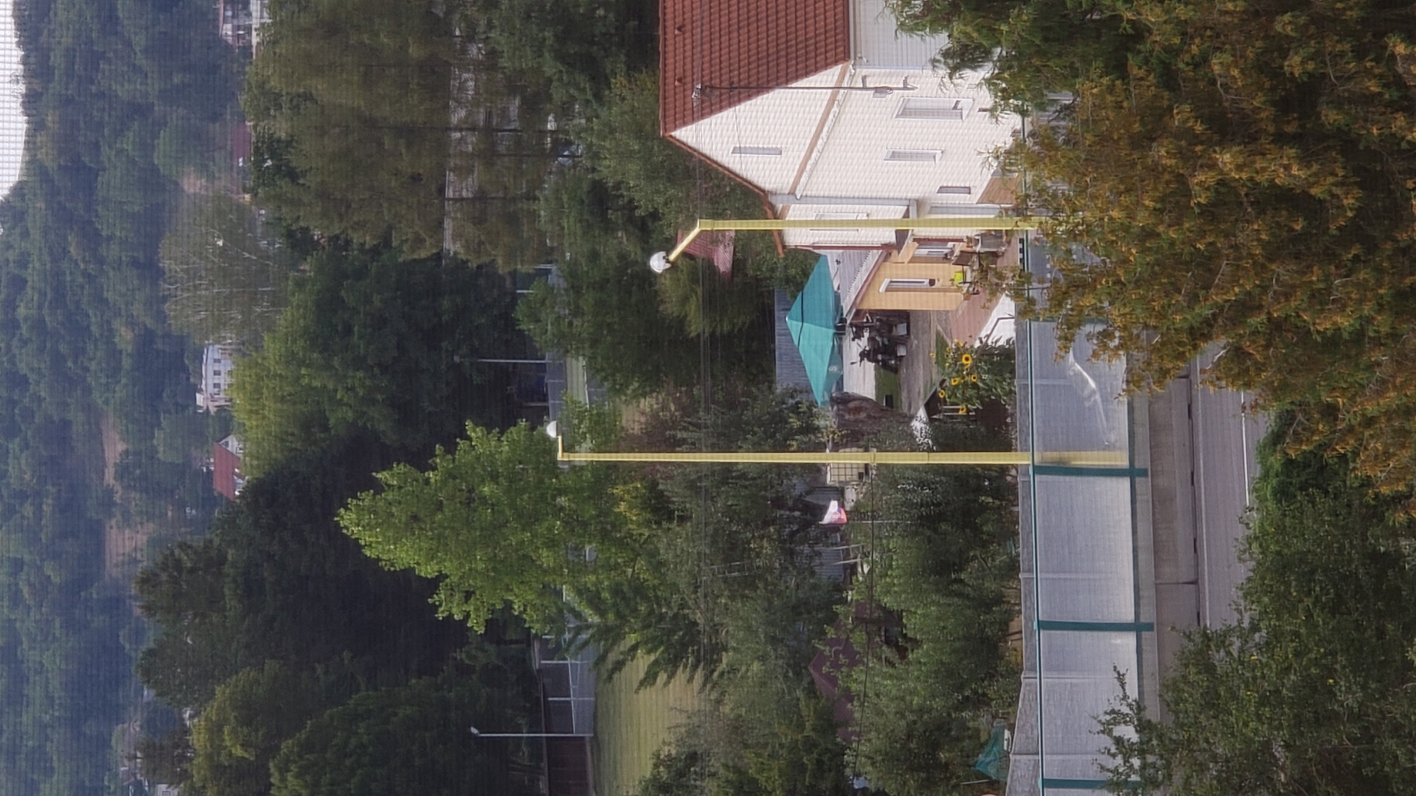 The image depicts an aerial view of a suburban or semi-rural area, likely on the outskirts of Prešov, Slovakia, judging by the presence of a road barrier commonly found along highways. In the foreground, a concrete and translucent barrier with green trim runs horizontally across the image, separating a road from the area behind it.
Behind the barrier, several elements are visible. Two tall, slender yellow lampposts with white spherical lights stand prominently. To the left of the lampposts, a cluster of trees and smaller structures suggest residential or recreational use. A green umbrella covers an outdoor seating area, with a few motorcycles parked nearby. Sunflowers are visible in front of a small building.
Further back, a white building with a red-tiled roof is situated. The building has multiple windows and appears to be a house or a small commercial establishment. Adjacent to this building, more trees and vegetation form a dense backdrop.
In the background, a lush, tree-covered hillside rises, dotted with a few buildings, some with red roofs, suggesting a residential area built into the slope. The overall impression is a landscape that blends natural elements with human habitation, characteristic of a town or city extending into its surrounding environment. The lighting suggests it is daytime, possibly late afternoon, given the shadows. No individuals are clearly discernible in the image.