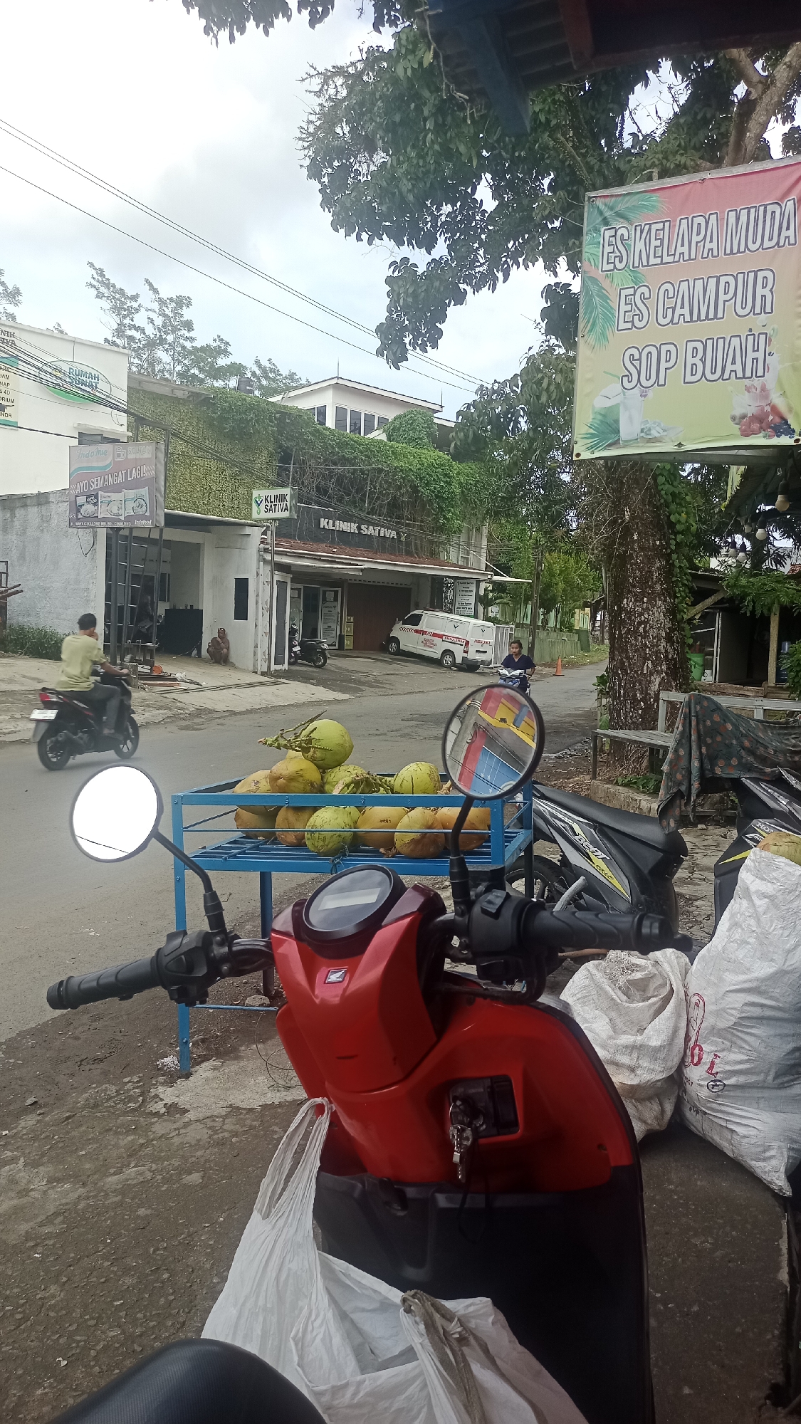 This is an outdoor, eye-level, medium shot image taken from behind a red scooter, looking towards a street scene in Cikalong, Indonesia. The foreground is dominated by the red scooter, with its handlebars, front panel, and a white plastic bag hanging from it visible. A portion of another scooter, black and yellow, is also seen to the right.
In the mid-ground, a blue metal stand is laden with a pile of young coconuts. Behind the coconuts, a road stretches with some pavement and a dirt shoulder. A few motorcycles are parked or in motion. One red scooter with a rider in a light green shirt is heading away from the camera on the left. Another scooter, a black and yellow model, is being ridden by a person with dark hair towards the right. Further back, a white ambulance with "AMBULANCE" written on its side is parked in front of a building.
The background consists of buildings and lush green trees. A banner hangs on a tree to the right, displaying text in Indonesian that appears to be an advertisement for drinks like "ES KELAPA MUDA" (young coconut ice), "ES CAMPUR" (mixed ice), and "SOP BUAH" (fruit soup). Buildings with various signs, including "KLINIK SATIVA" (Sativa Clinic), are visible along the street. Power lines crisscross the sky, which is overcast with white clouds. The overall impression is of a casual street-side scene, possibly a market or roadside stall, in a town or city.