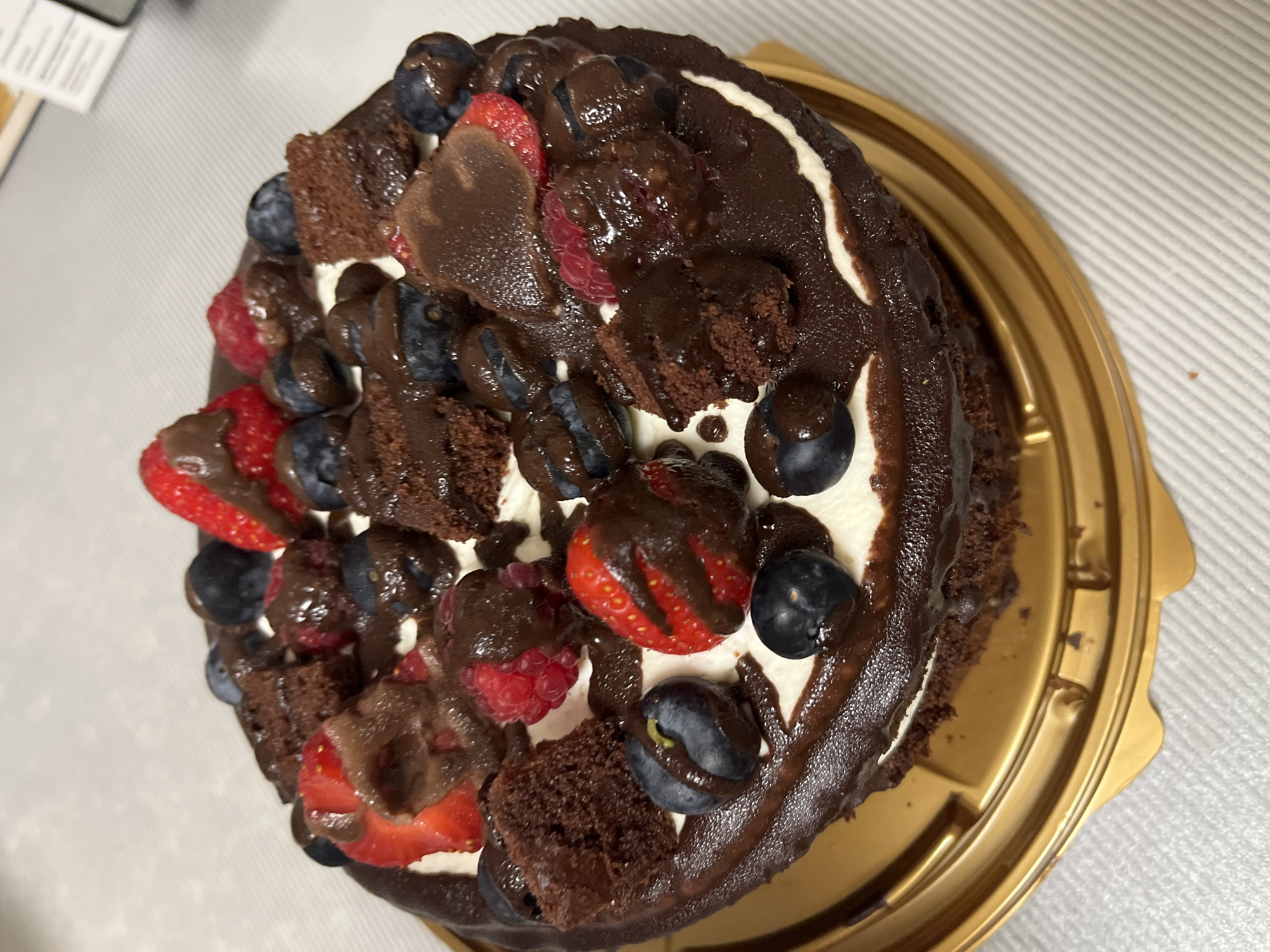 A chocolate cake topped with whipped cream, fresh berries, and chunks of brownie is presented on a gold plastic cake stand. The cake features a dark chocolate frosting and is drizzled with melted chocolate. Visible berries include strawberries, raspberries, and blueberries. The cake is situated on a light-colored, textured surface. The background is blurred, but appears to be indoors, with some white elements that could be a wall or packaging. There are no people visible in the image. The location is not specified.