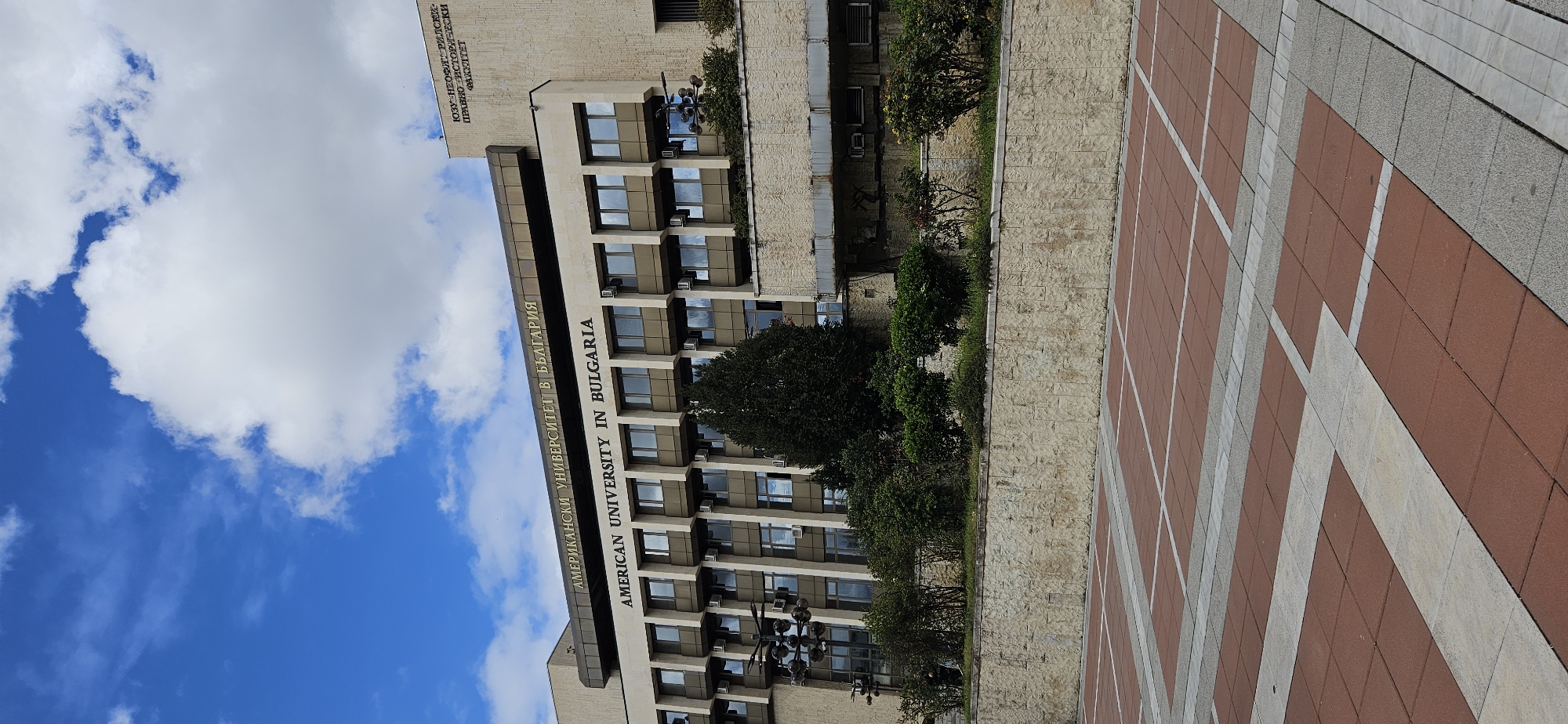 The image depicts a building with the inscription "AMERICAN UNIVERSITY IN BULGARIA" in both English and Bulgarian. The building has multiple floors with windows and is located in Blagoevgrad, Bulgaria. The architecture appears modern, and there is a paved area in front of the building. The sky is visible with clouds, suggesting daylight. There is also greenery, including trees and bushes, around the building. The foreground shows a tiled pattern on the ground.