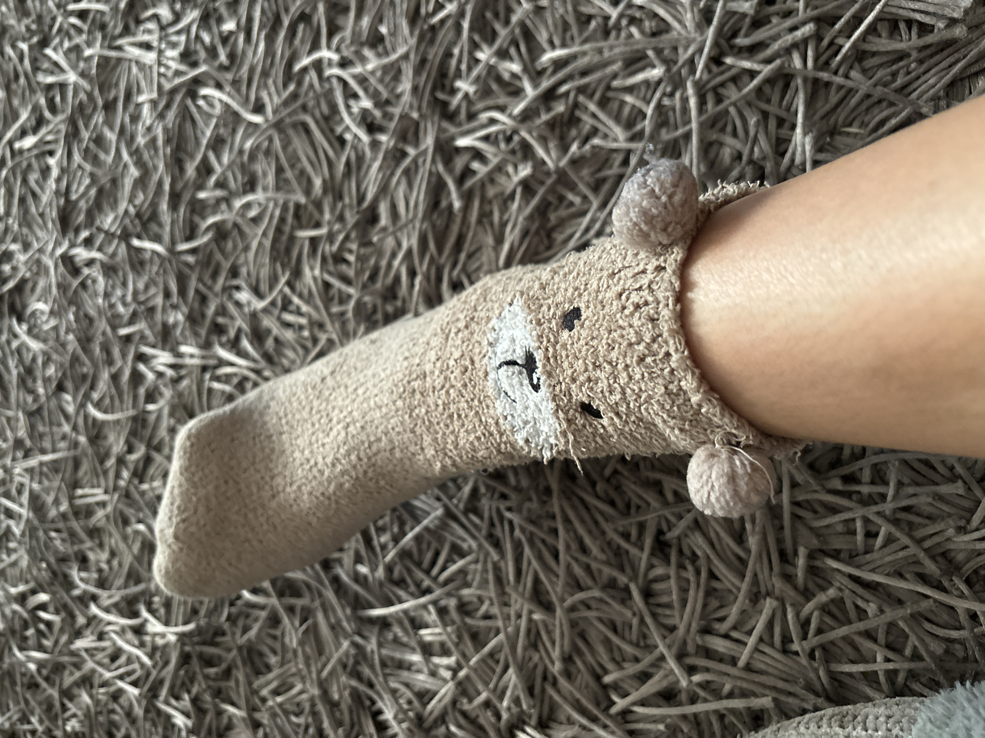 The image shows a person's lower leg and foot wearing a novelty sock designed to resemble a bear. The sock is light brown and features a bear's face on the ankle area, with two fuzzy pom-poms attached near the top edge. The person is standing on a gray, shag-style rug. The location in the city of Budva, Montenegro is not known.