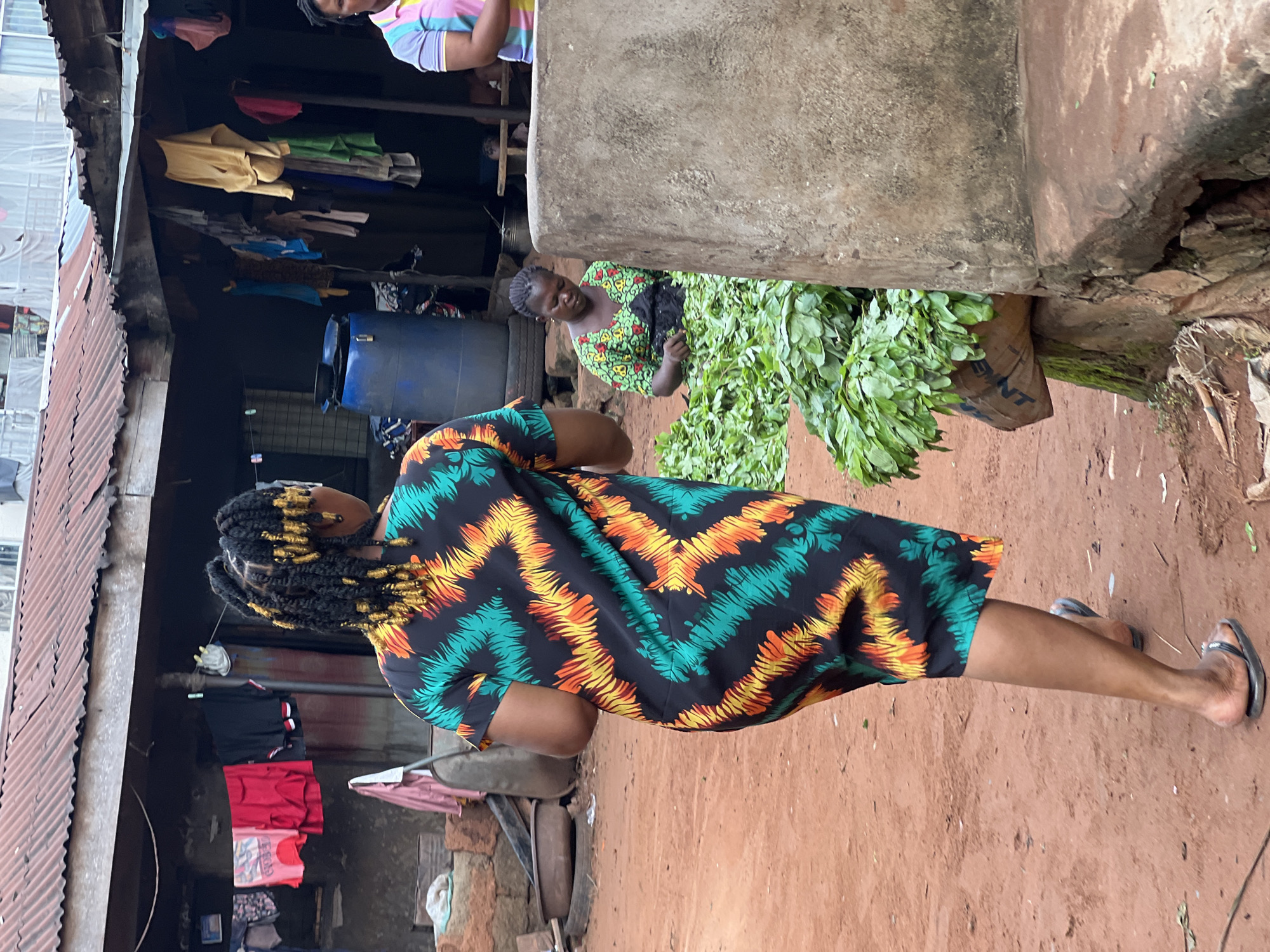 In Nkpor, Nigeria, a woman walks past a stall at a marketplace. She is wearing a black dress with a zigzag pattern of green, orange, and yellow. Her hair is braided with gold accents. In the background, another woman is seated, sorting through a large pile of green leafy vegetables. The marketplace appears to be outdoors, with corrugated metal roofing and dirt ground. Laundry hangs on a line in the background, and a large blue plastic barrel is visible. The scene suggests daily commerce and community life in this Nigerian city.
