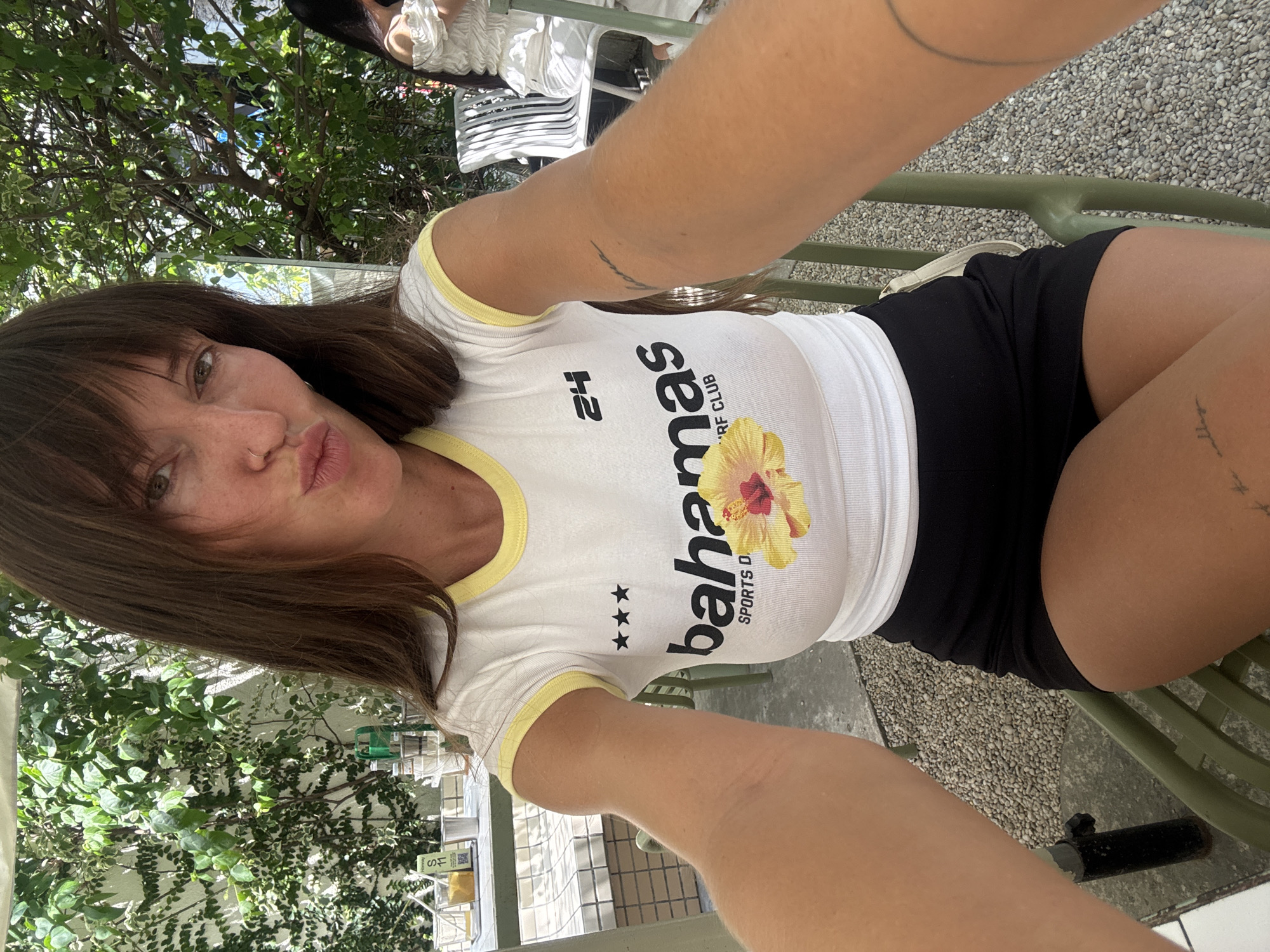 A person with brown hair and bangs is taking a selfie. They are wearing a white t-shirt with yellow trim that says "Bahamas Sports D. RF Club" and black shorts. They are sitting on a green metal bench outside, with green foliage in the background and gravel on the ground. Based on the style of furniture and background elements, the location could be Đà Nẵng, Vietnam.