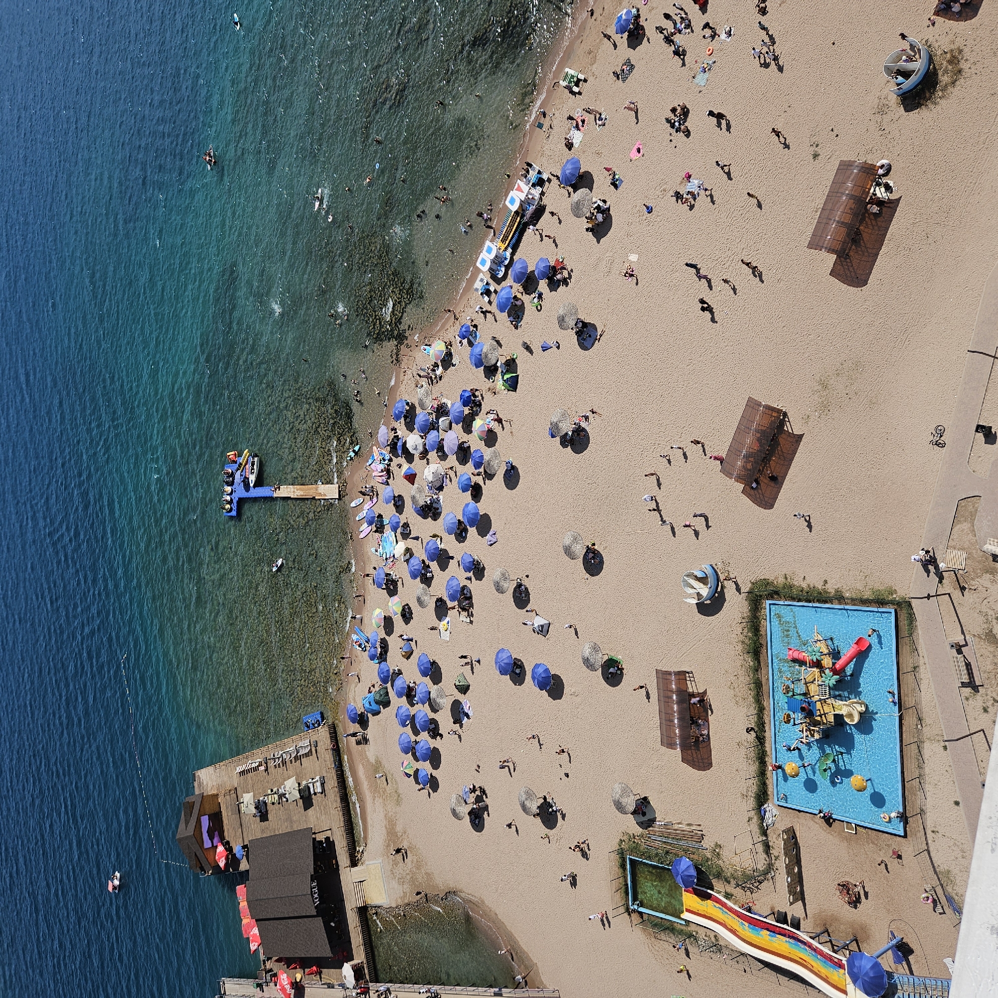 This is an aerial, full-frame, high-angle shot of a beach in Bosteri, Kyrgyzstan, along the shore of Lake Issyk-Kul. The water is a vibrant turquoise, with clear shallows revealing the rocky lakebed near the shore. The sand on the beach is a light tan color and is dotted with numerous blue and grey umbrellas, along with a few green tents, providing shade for beachgoers.
There are many people on the beach, some walking, some sitting or lying under umbrellas, and some in the water. Several watercraft are visible: a small white boat with an orange top is docked at a wooden pier with a small structure on it, and a yellow and blue catamaran-like vessel is also moored near the pier. A separate, longer wooden pier extends into the water with a blue platform at its end, occupied by several individuals.
Further inland, a children's water play area is visible with a blue swimming pool, a red and yellow water slide, and a yellow water play structure with slides and palm tree decorations. Another smaller, rectangular pool is located to the left of the main water play area.
Scattered across the beach are various other objects, including lounge chairs, small tables, and some portable shaded structures. A bicycle is parked near a paved pathway in the lower right portion of the image. The overall scene depicts a busy recreational area with a clear focus on water activities and beach relaxation.